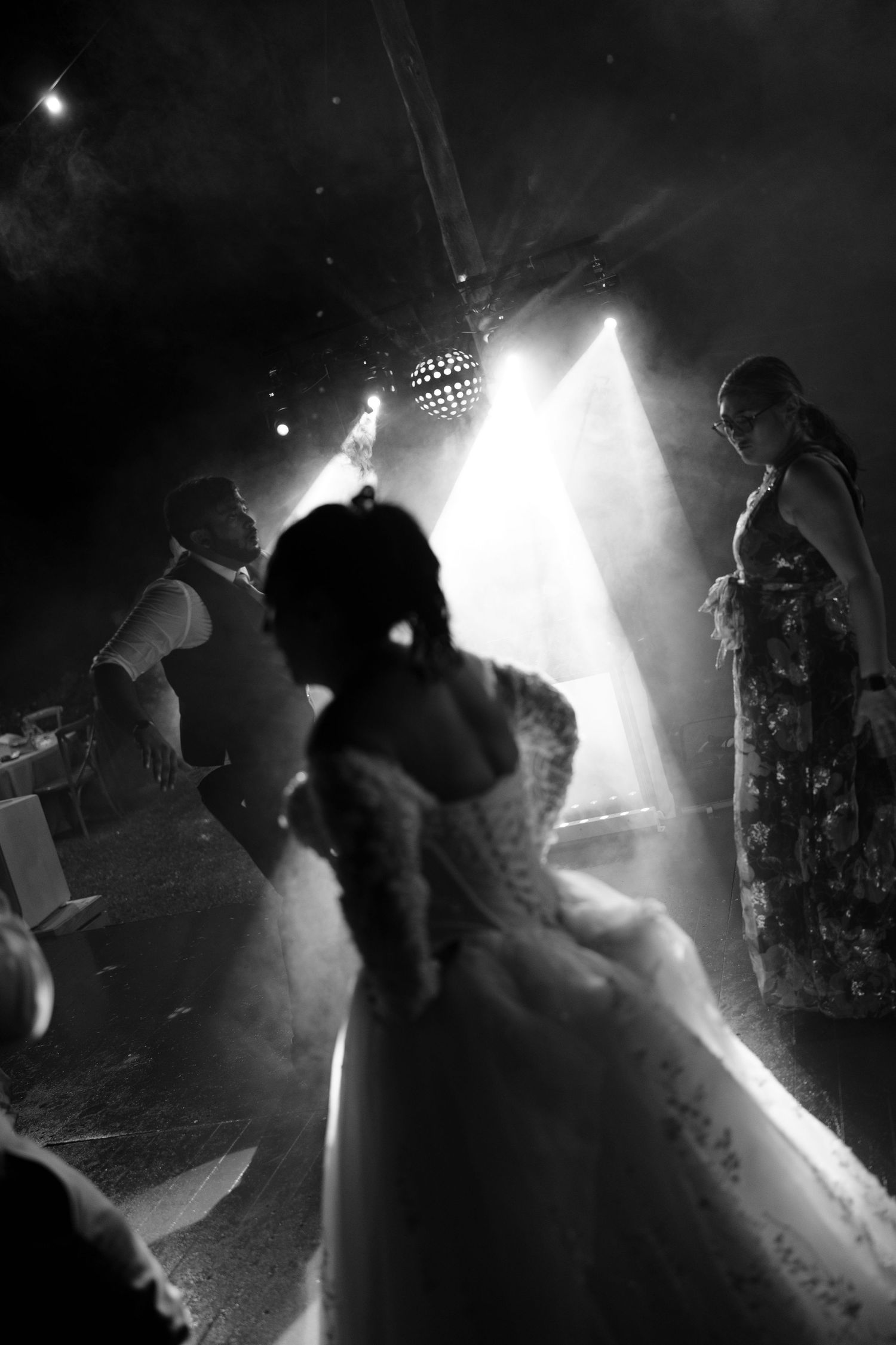 Black and white photo of wedding guests dancing under dramatic stage lights and haze, with a bride in a lace gown in the foreground.