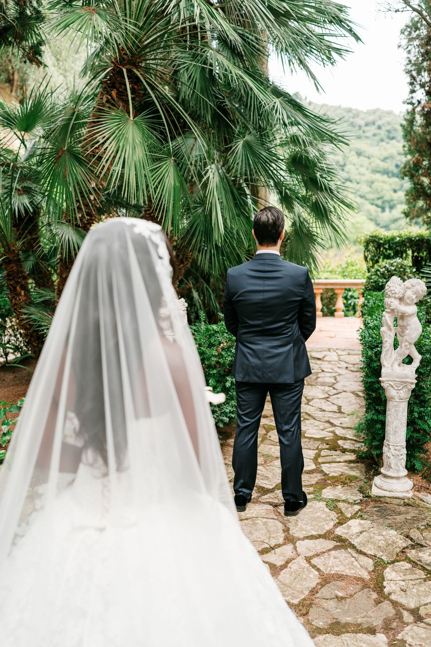Bride in a long lace veil and gown faces a groom in a dark suit on a stone garden path lined with palm trees and a cherub statue.