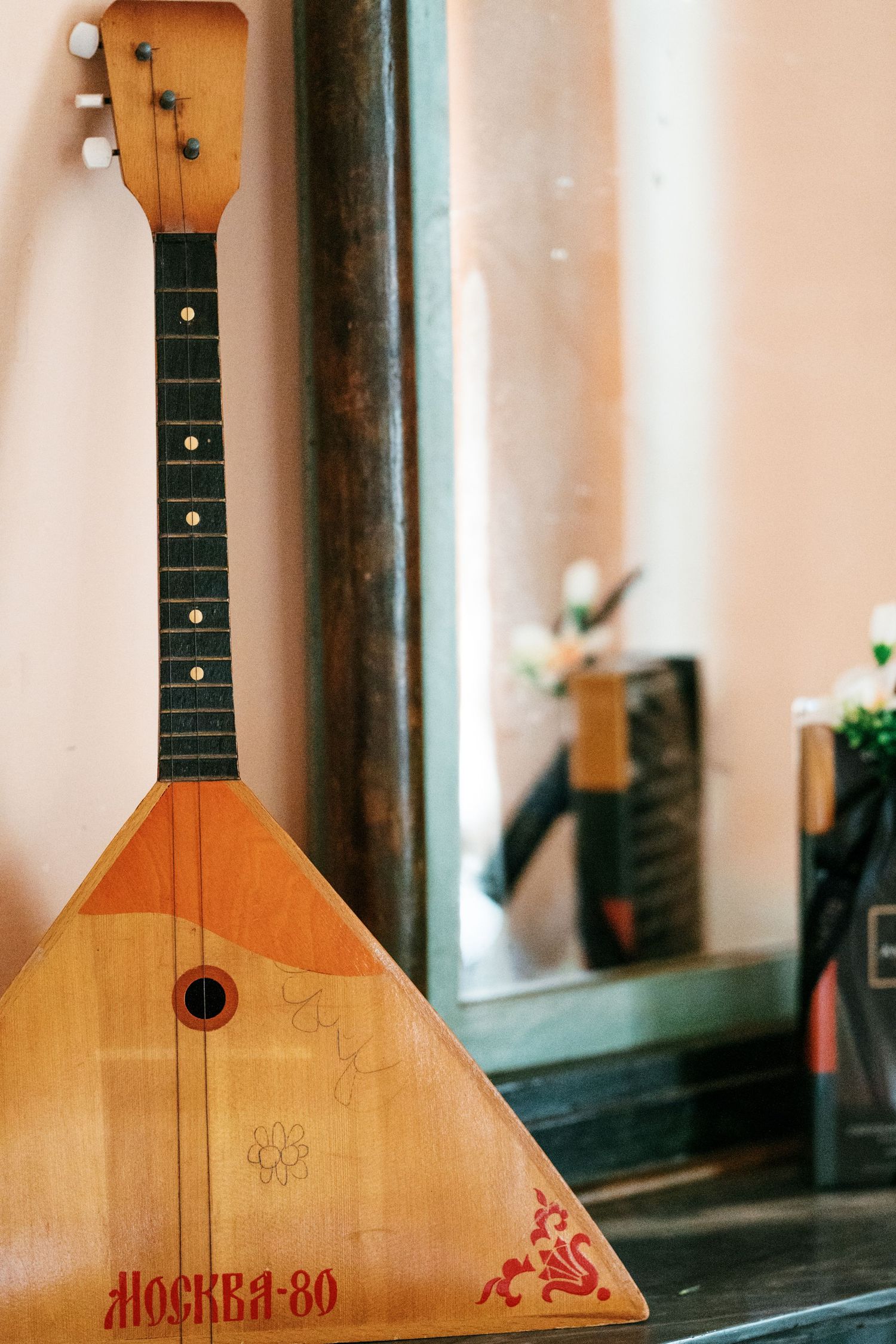 Wooden triangular balalaika with red Moscow-80 lettering and floral designs leans against a mirror reflecting flowers.