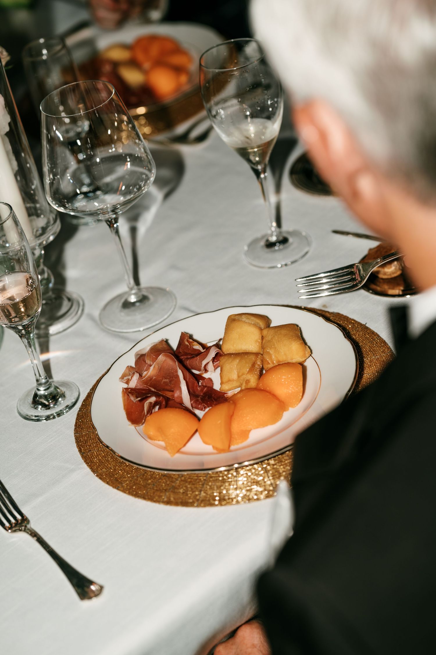 Elegant dinner table setting with prosciutto and cantaloupe appetizer on a gold glitter charger plate beside crystal wine glasses.