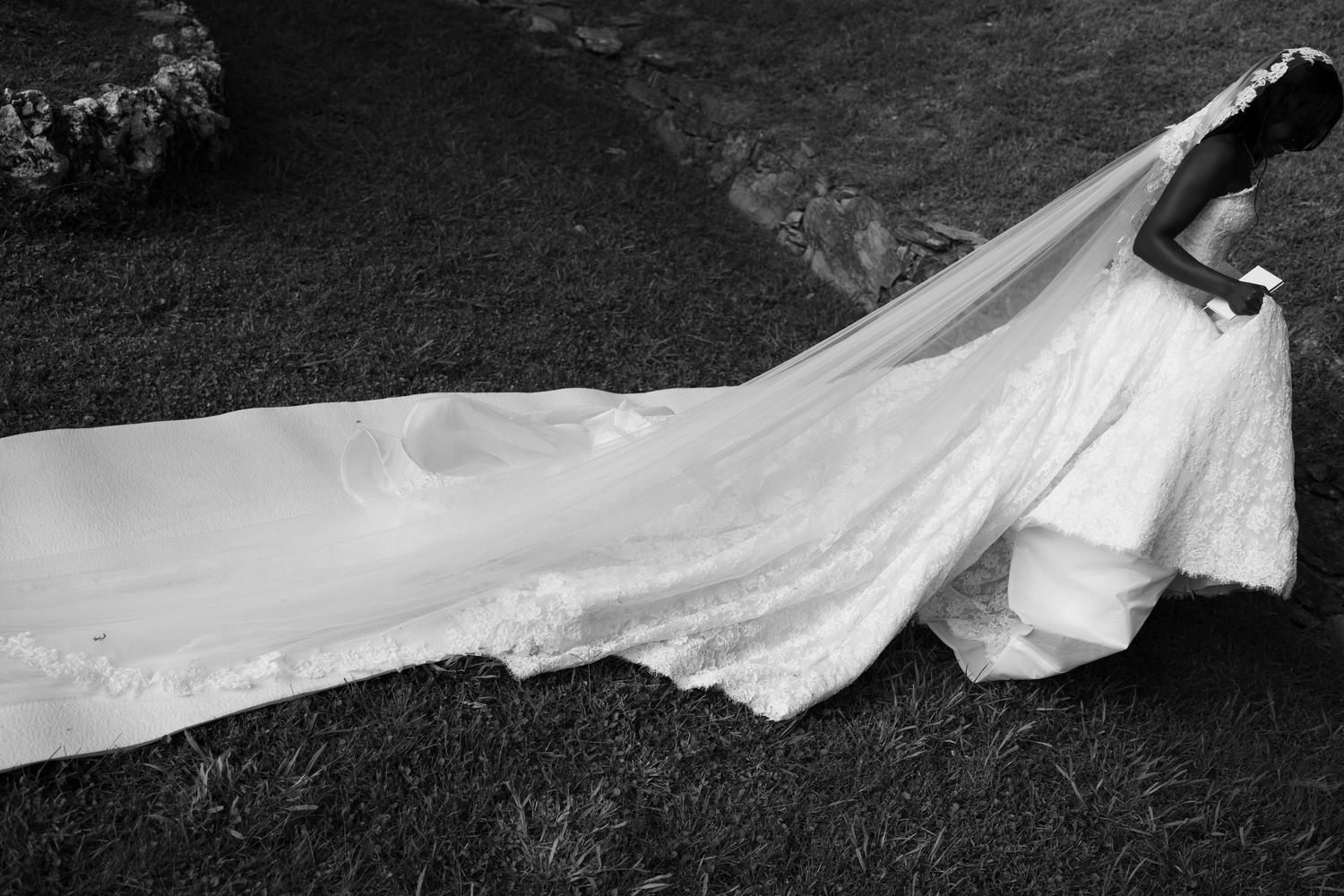 Black and white photo of a bride in a lace wedding gown with a long veil and train stretching across a grassy lawn.