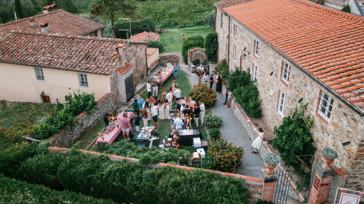 Wedding guests gather at long tables in a garden courtyard of a rustic Tuscan villa with terracotta roofs and manicured hedges.