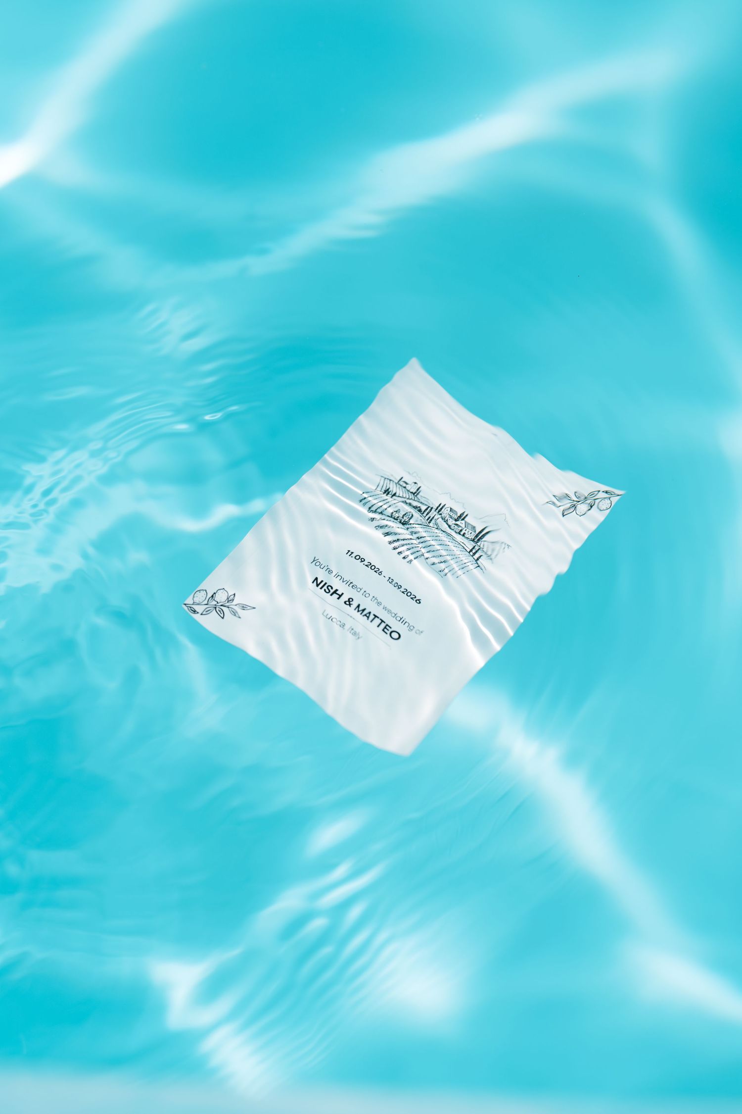 A white wedding invitation card floats on rippling turquoise pool water, with sunlight creating bright reflections around it.