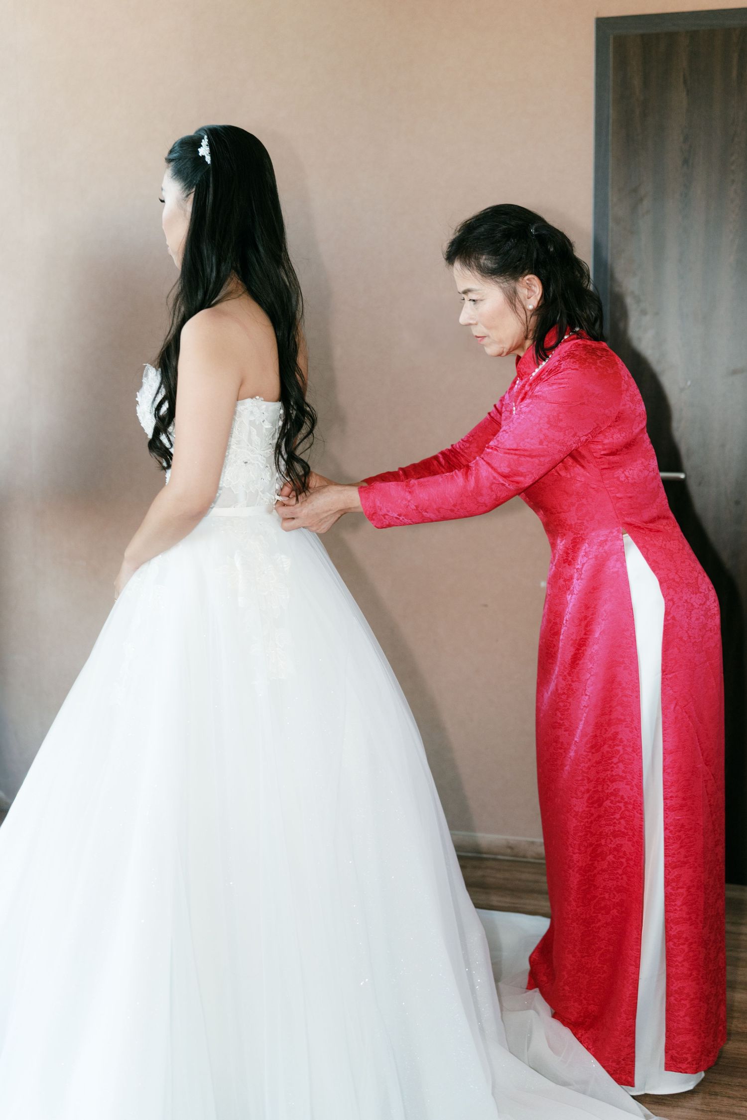 Mother in a red Vietnamese ao dai helps fasten the back of her daughters white wedding gown. Italy wedding photographer and filmmaker