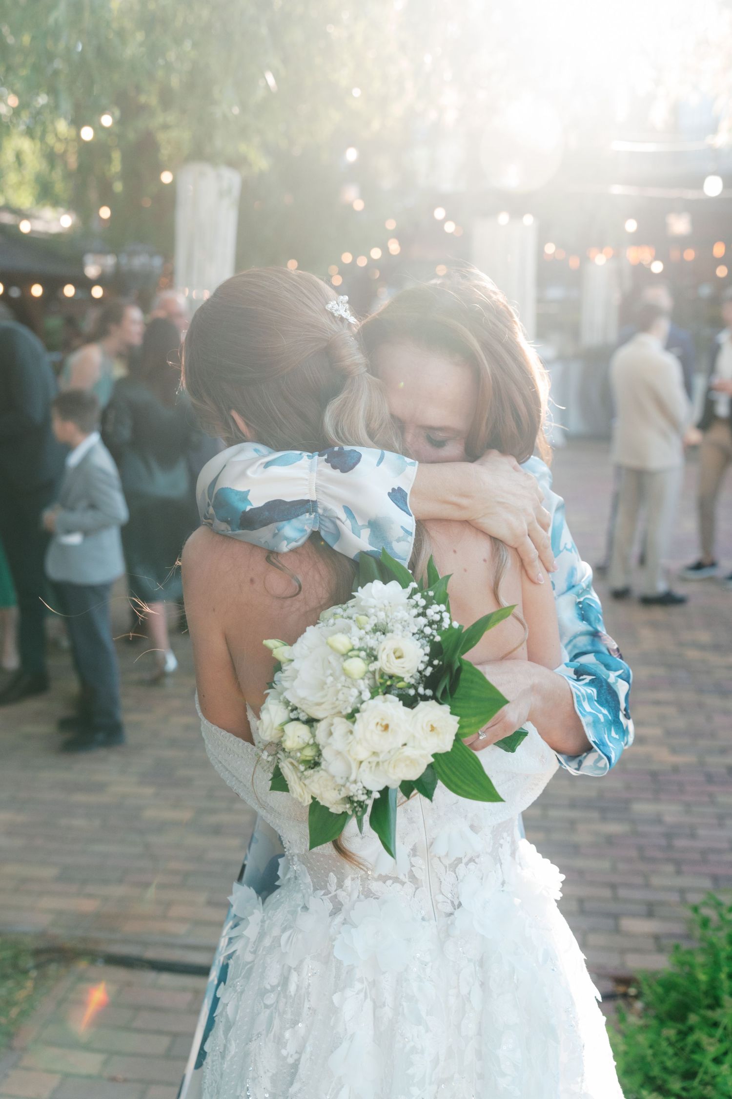 Bride holding white bouquet shares emotional hug with guest at outdoor wedding reception with string lights. Mallorca wedding photographer