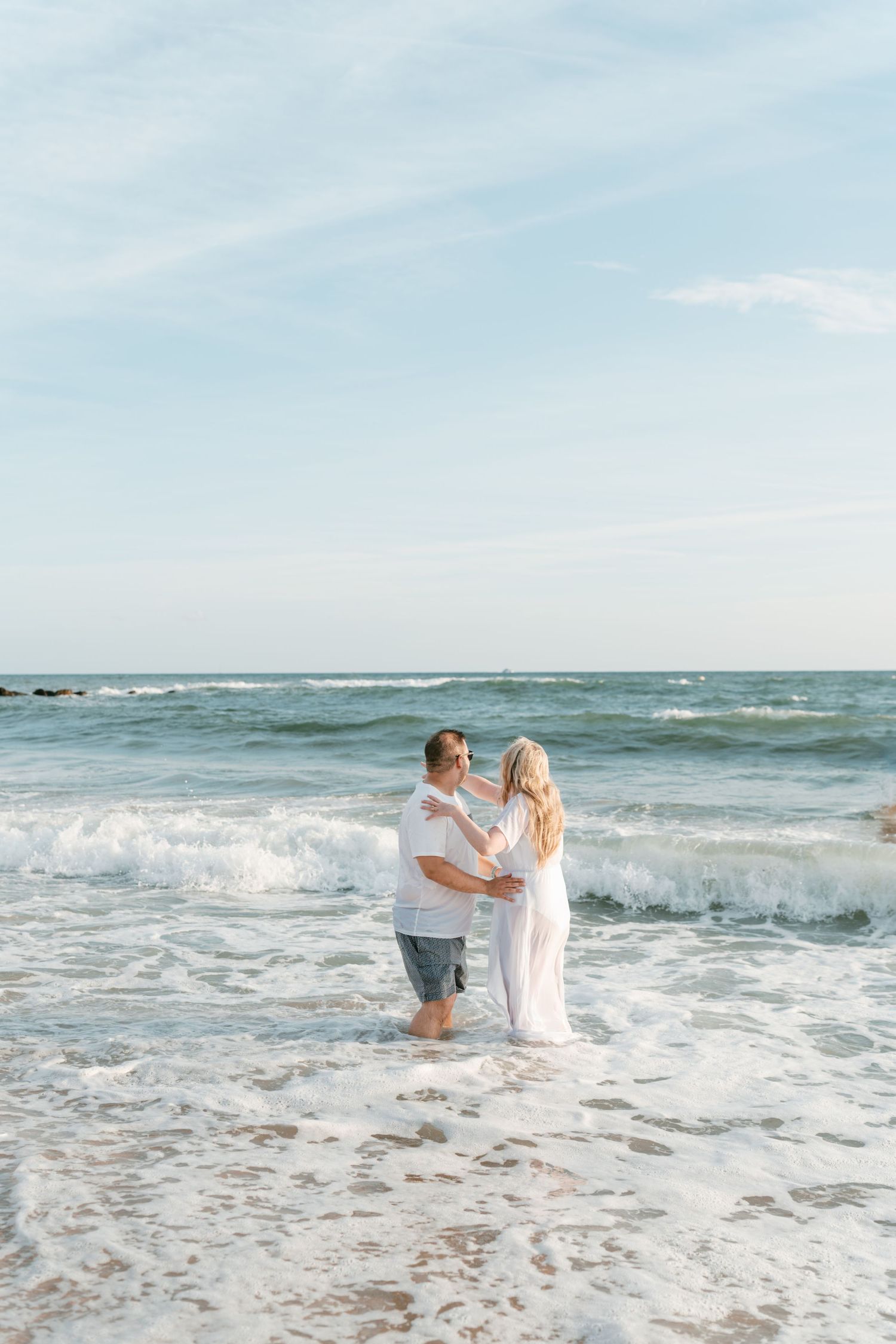 Couple dancing in ocean waves at the beach, woman in white dress and man in white shirt, romantic seaside moment. Mallorca wedding filmmaker