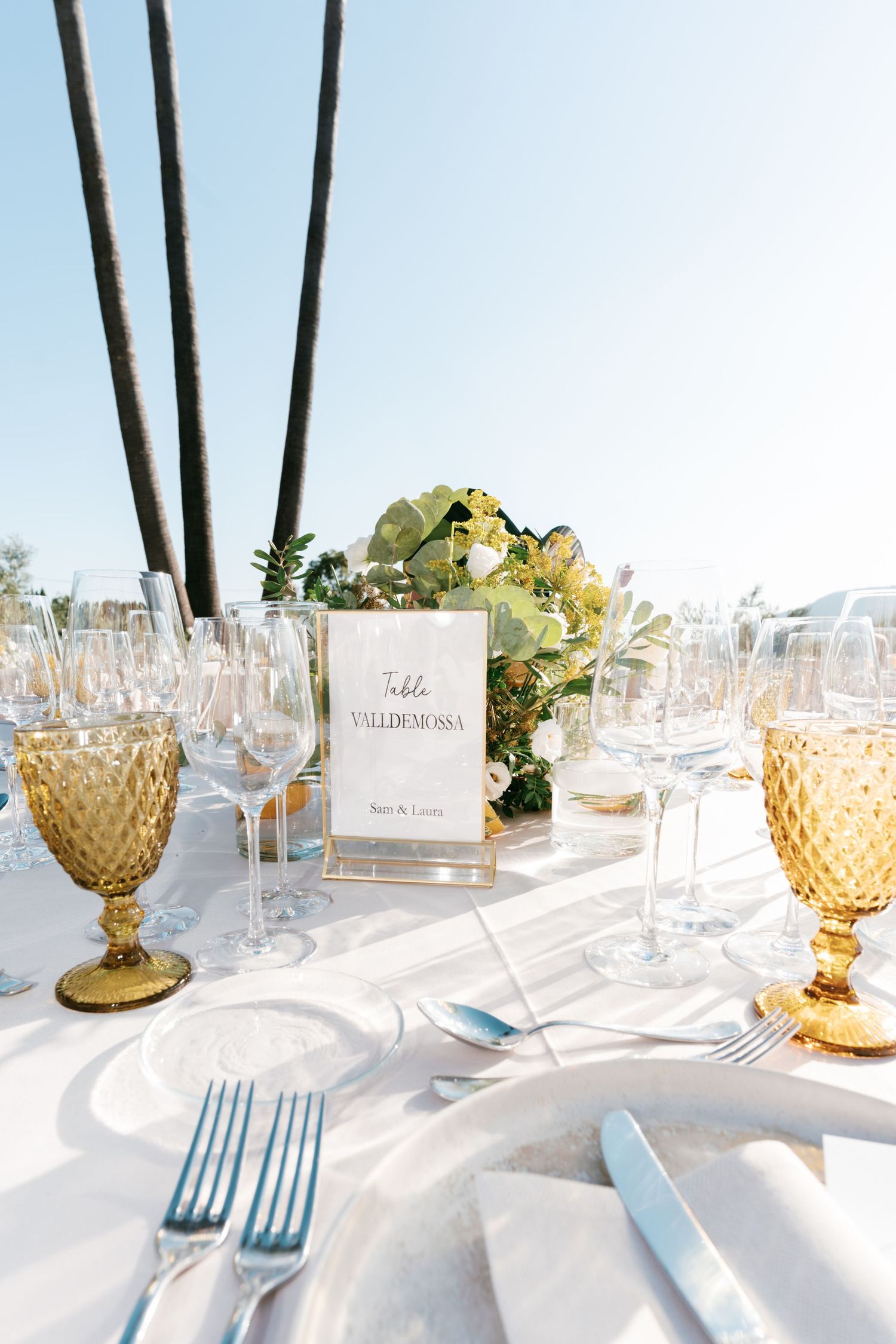 Elegant outdoor wedding table setting with amber glasses, white linens, floral centerpiece, and a Valldemossa table name card. Mallorca