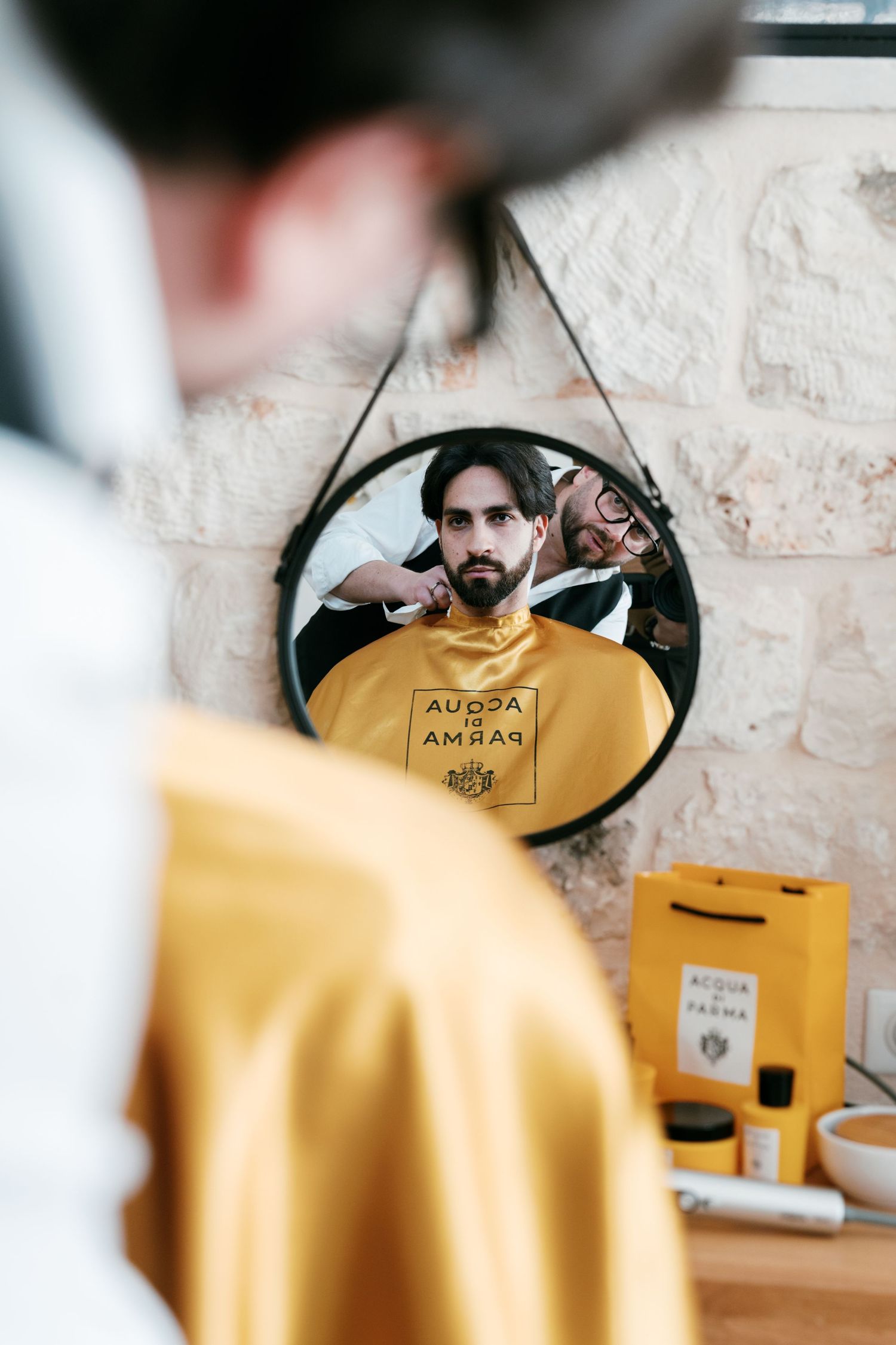 Groom getting groomed by a barber reflected in a round mirror with Acqua di Parma branding. Puglia wedding photographer, film