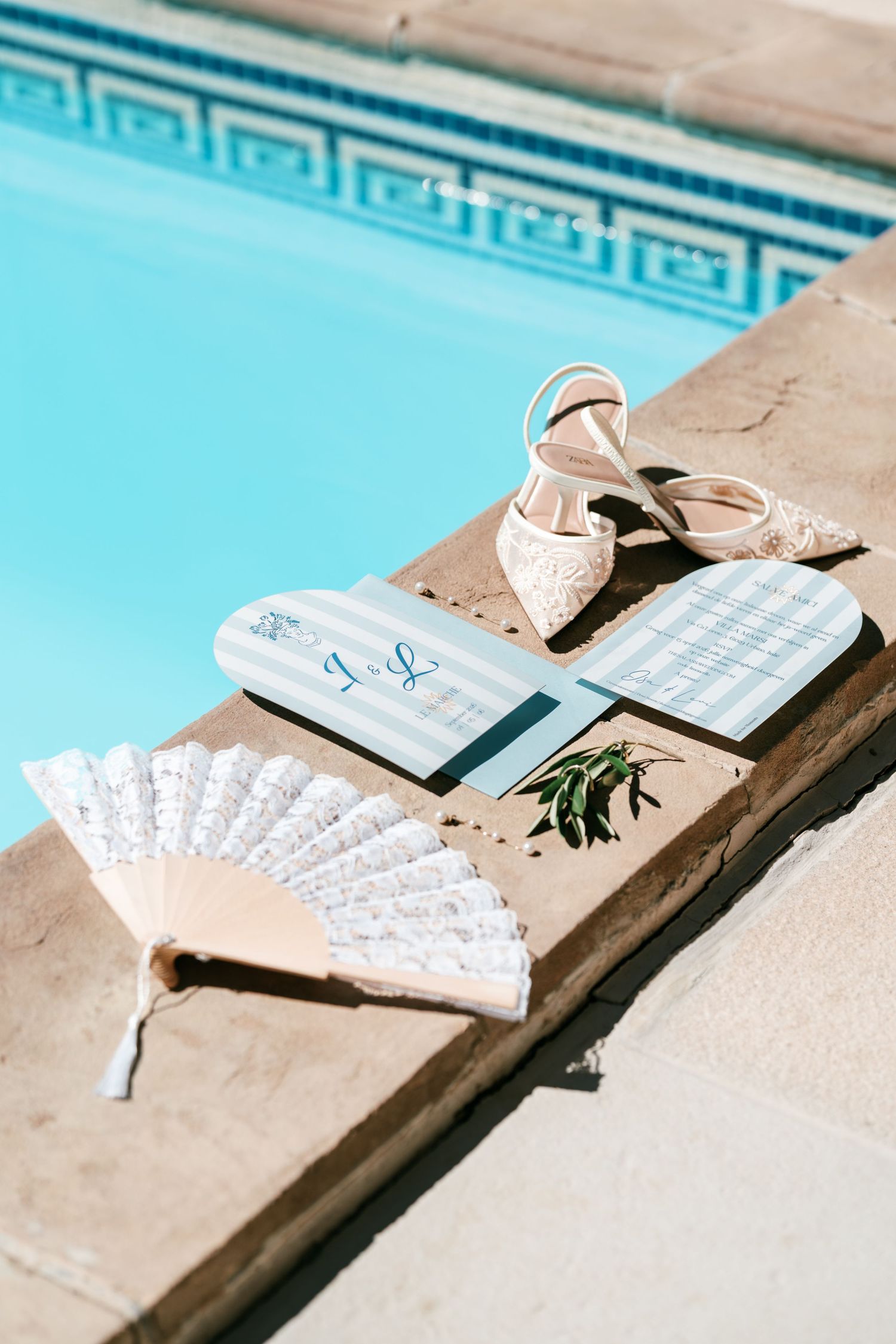 Elegant wedding flat lay with lace heels, hand fan, and striped programs by a blue pool.