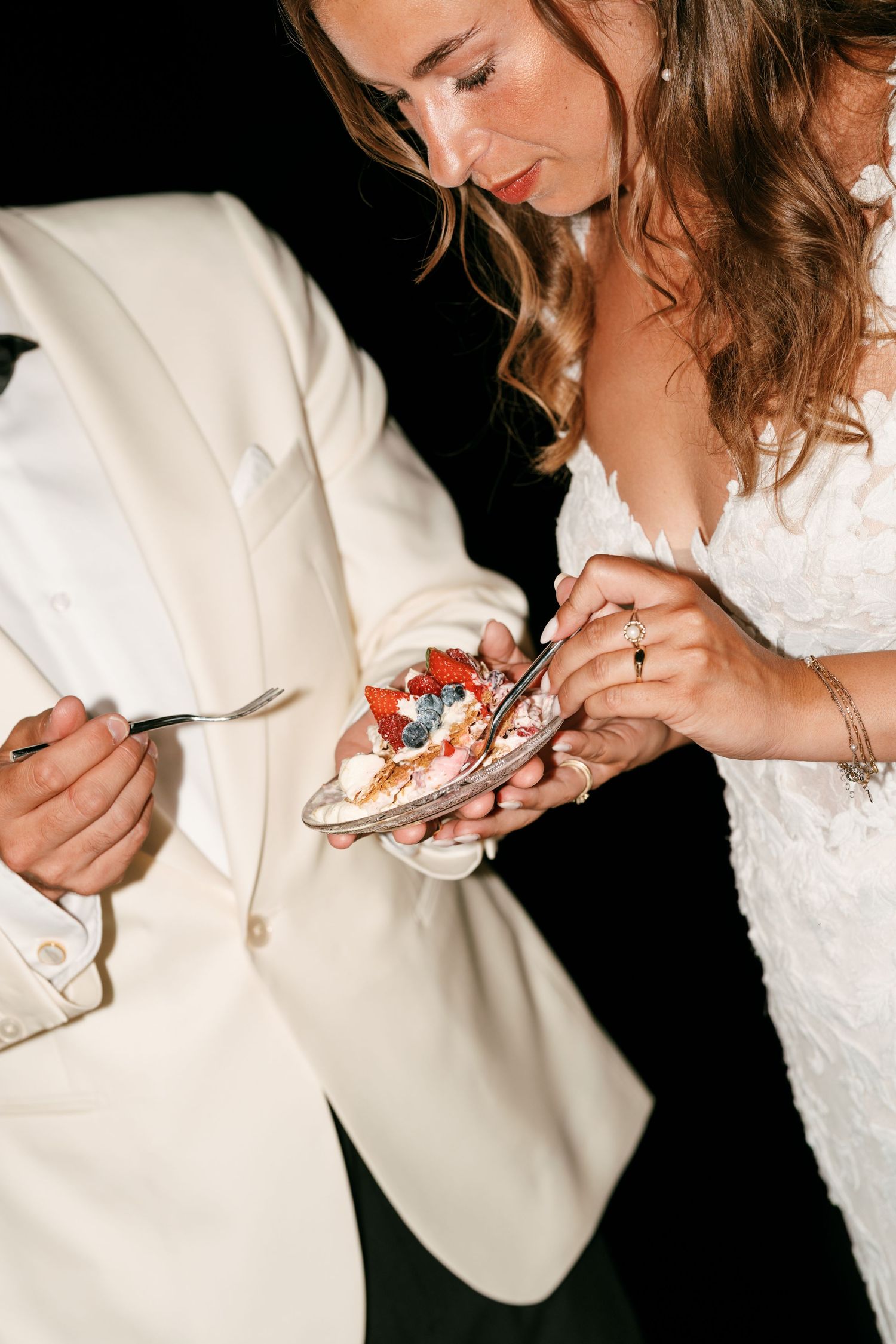 Bride and groom in elegant attire sharing a slice of wedding cake topped with fresh berries at their reception.