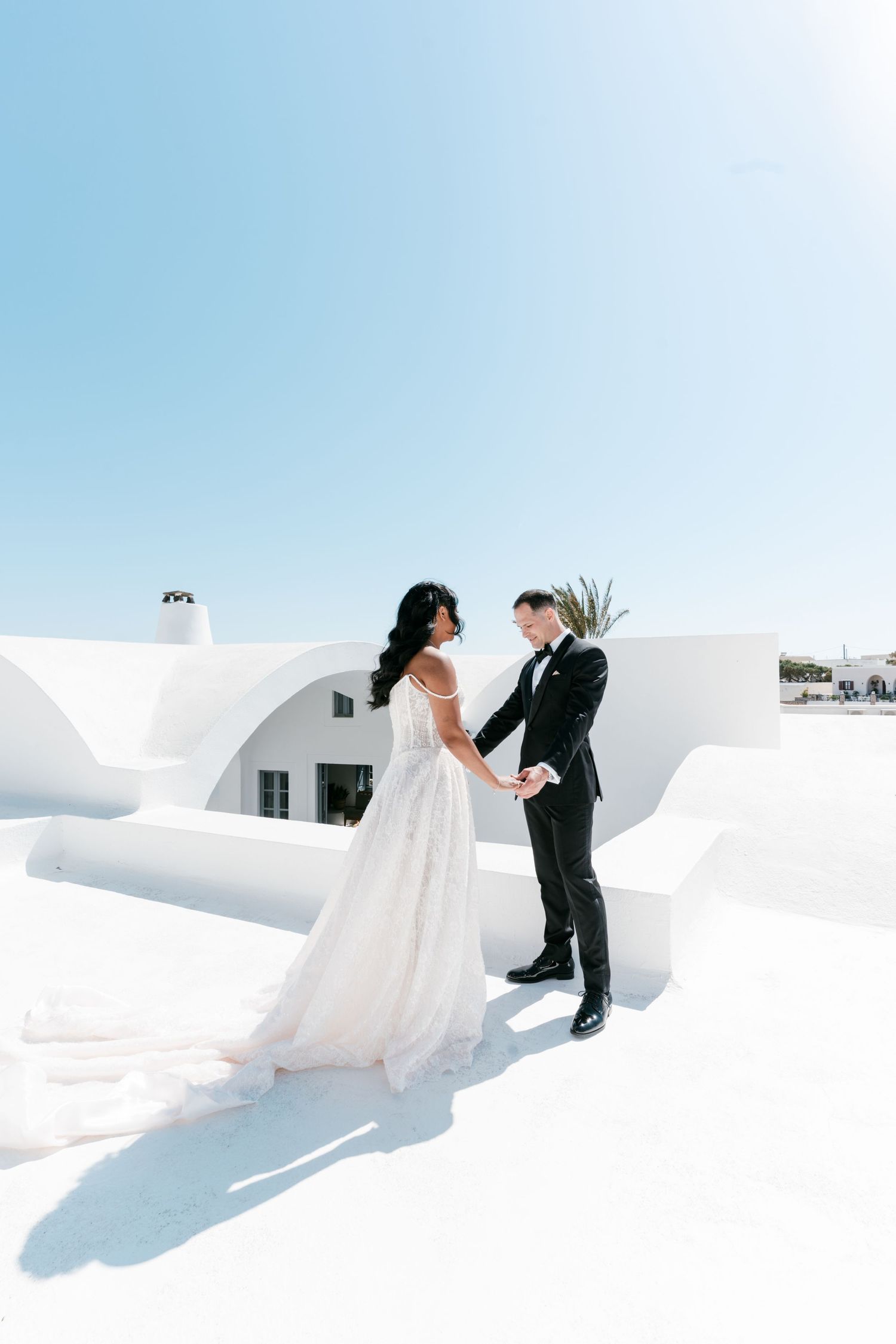 Bride and groom hold hands on white rooftop in Santorini, Greece, under a bright blue sky.