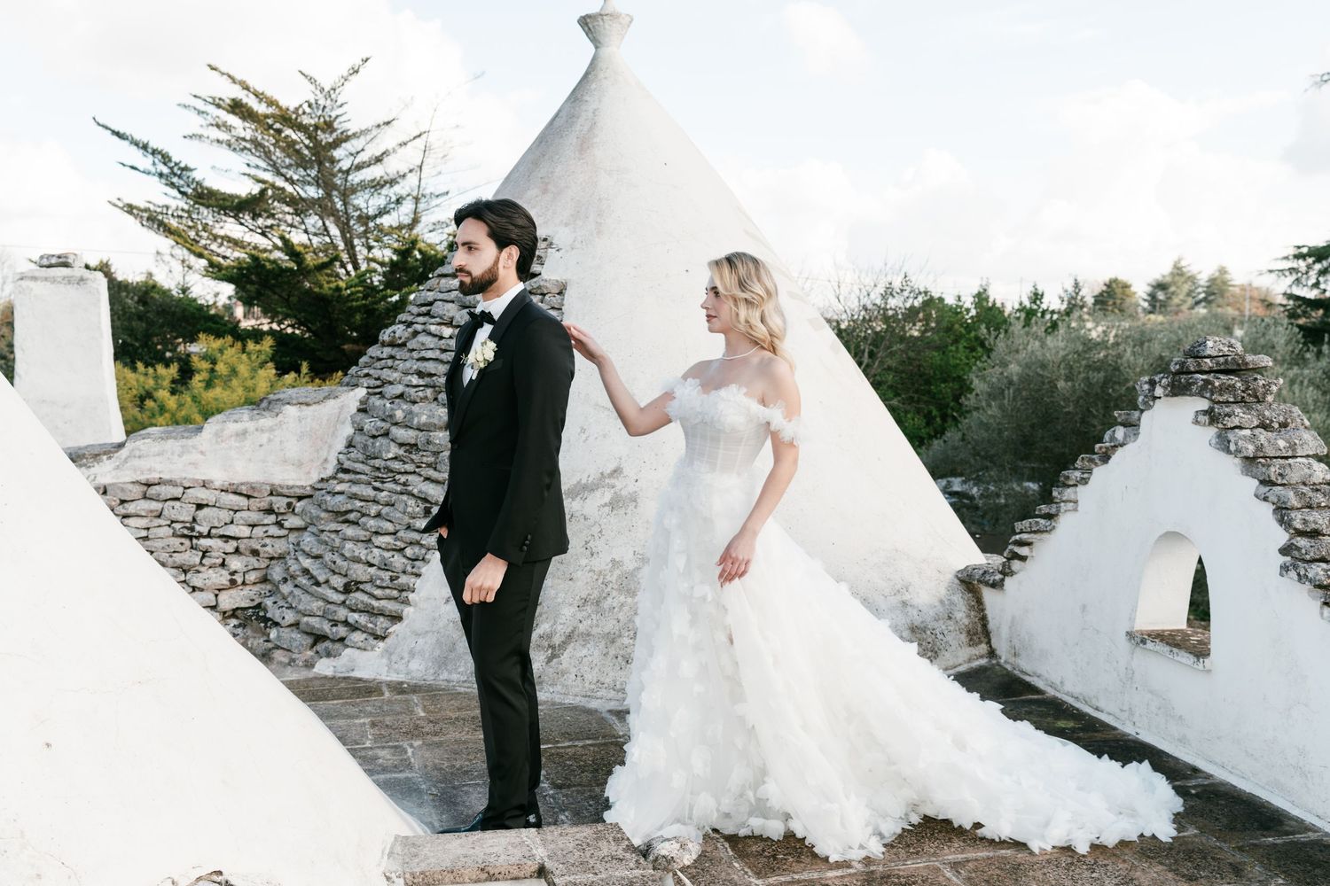 Bride in strapless ruffled gown and groom in black tuxedo pose near white trulli stone buildings at outdoor wedding.