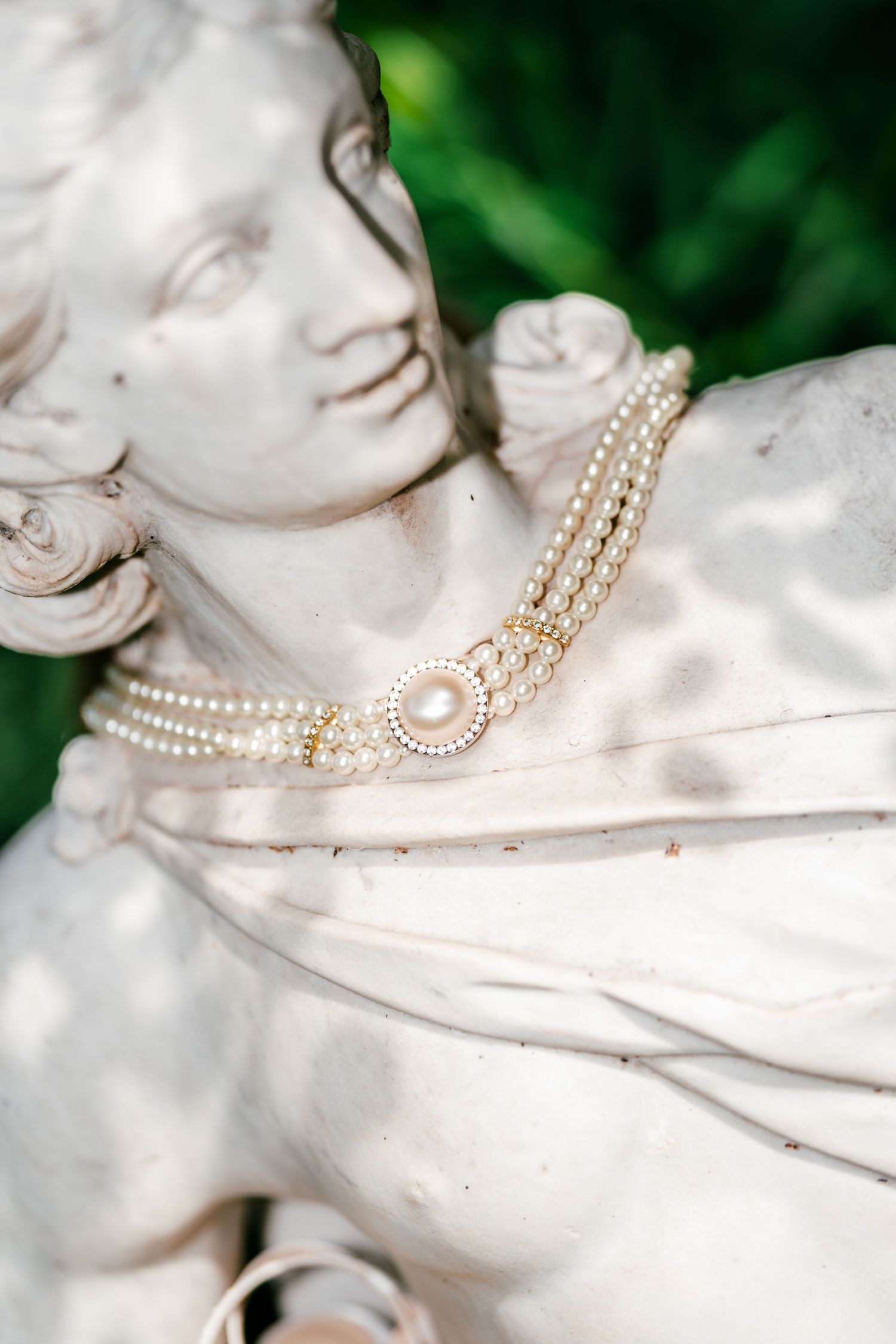 White stone statue of a young woman wearing a multi-strand pearl necklace with a large pearl and crystal clasp, set against green foliage.