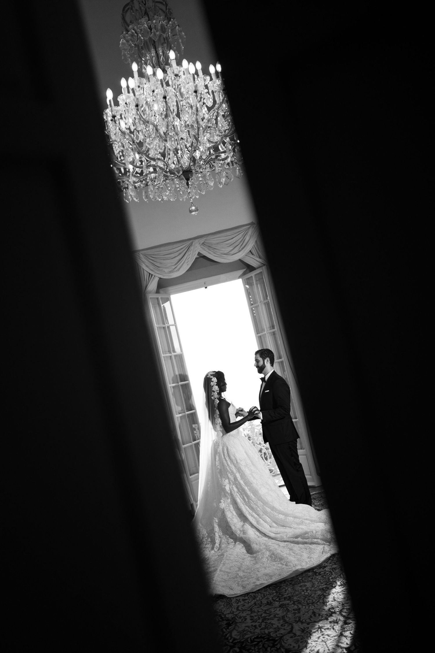Black and white photo of a bride and groom holding hands by a tall window, framed by a crystal chandelier overhead.