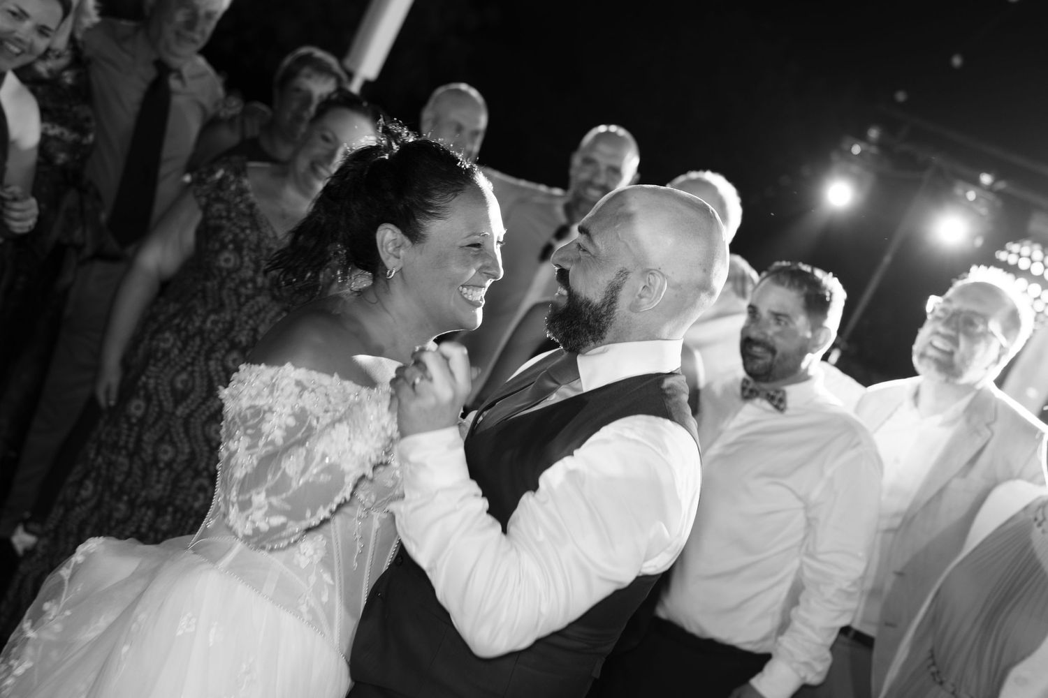 A smiling bride and groom celebrate with joyful wedding guests gathered around them on the dance floor.