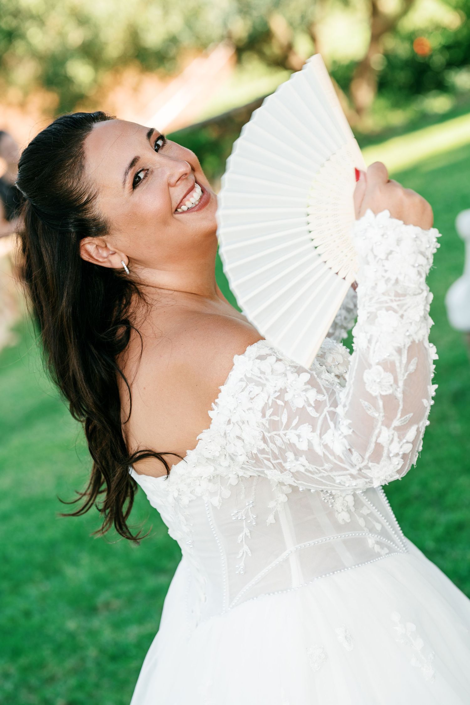 Smiling bride in an off-shoulder white lace wedding gown holds a white folding fan beside her face on a sunny green lawn.