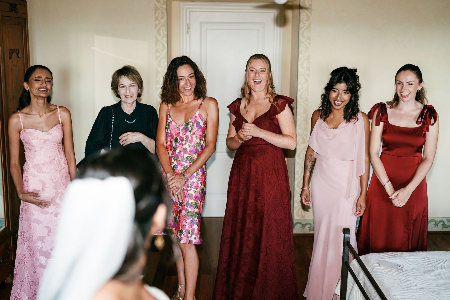 Six women in pink and burgundy dresses stand smiling in a bright room, looking toward a bride in a white veil in the foreground.