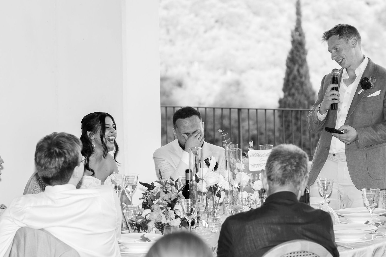 Wedding guest gives a speech with a microphone as the bride laughs and the groom covers his face at a reception table with flowers.