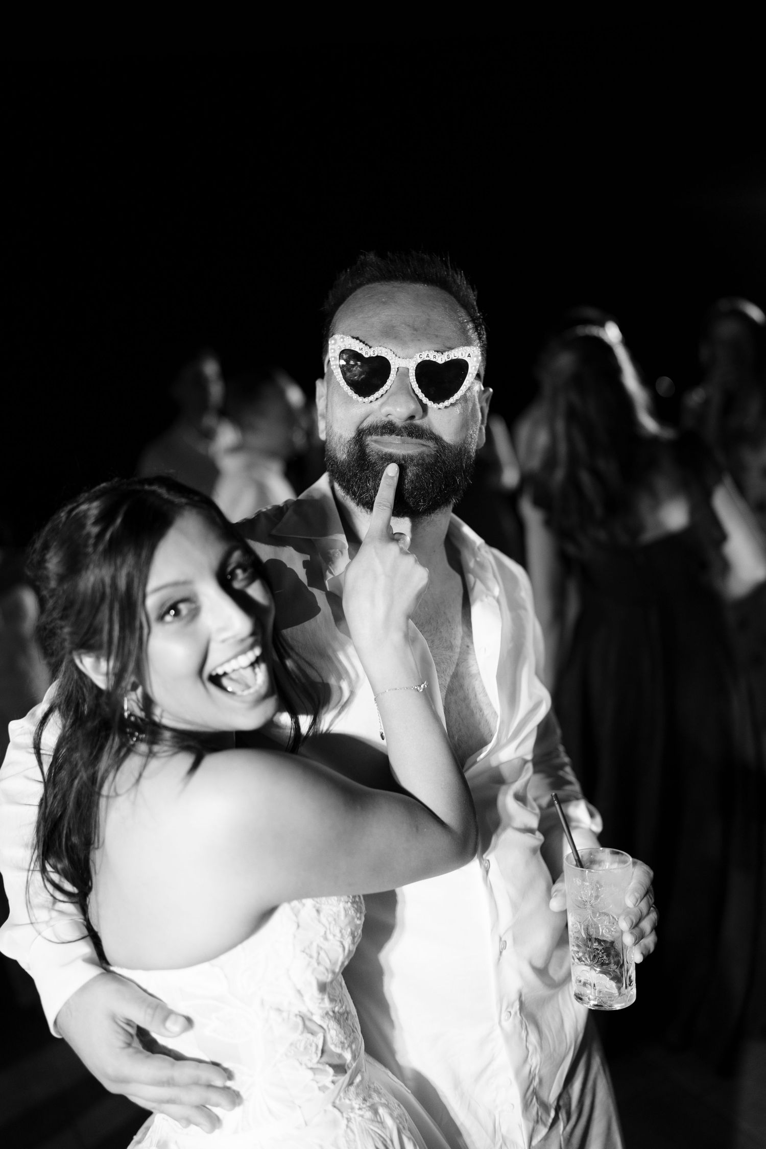 Black and white photo of a laughing bride in a lace gown posing with a bearded man in heart-shaped sunglasses at a wedding party.
