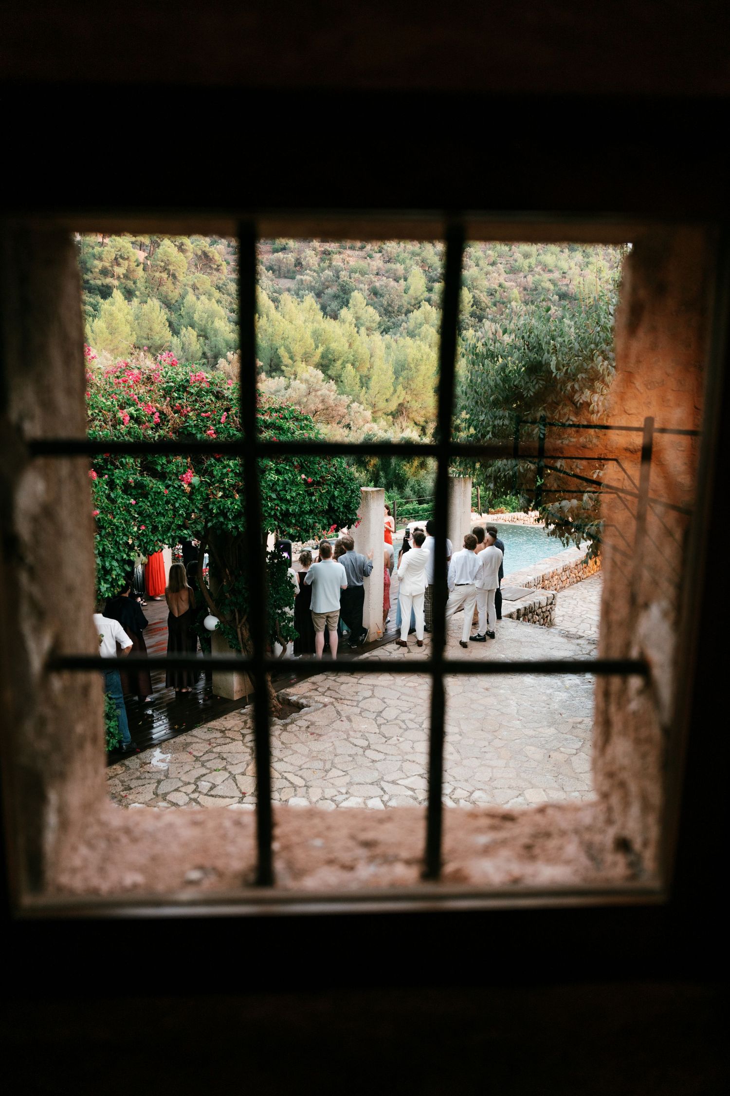 View through a barred stone window of wedding guests gathering on a patio beside a pool, with lush green hills and pink flowers beyond.
