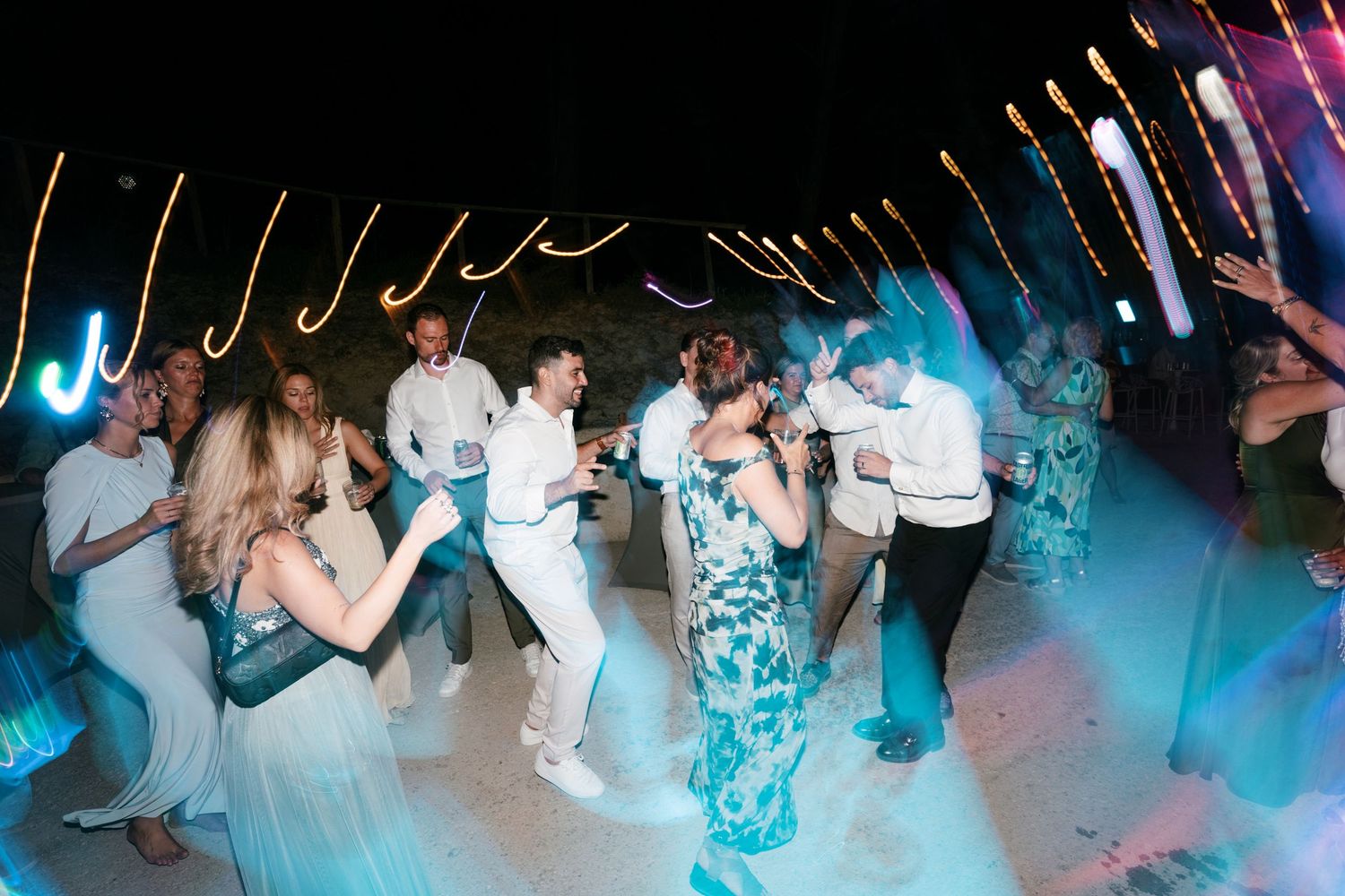 Guests dancing energetically at a nighttime wedding reception with colorful lights and sparklers creating a festive atmosphere.