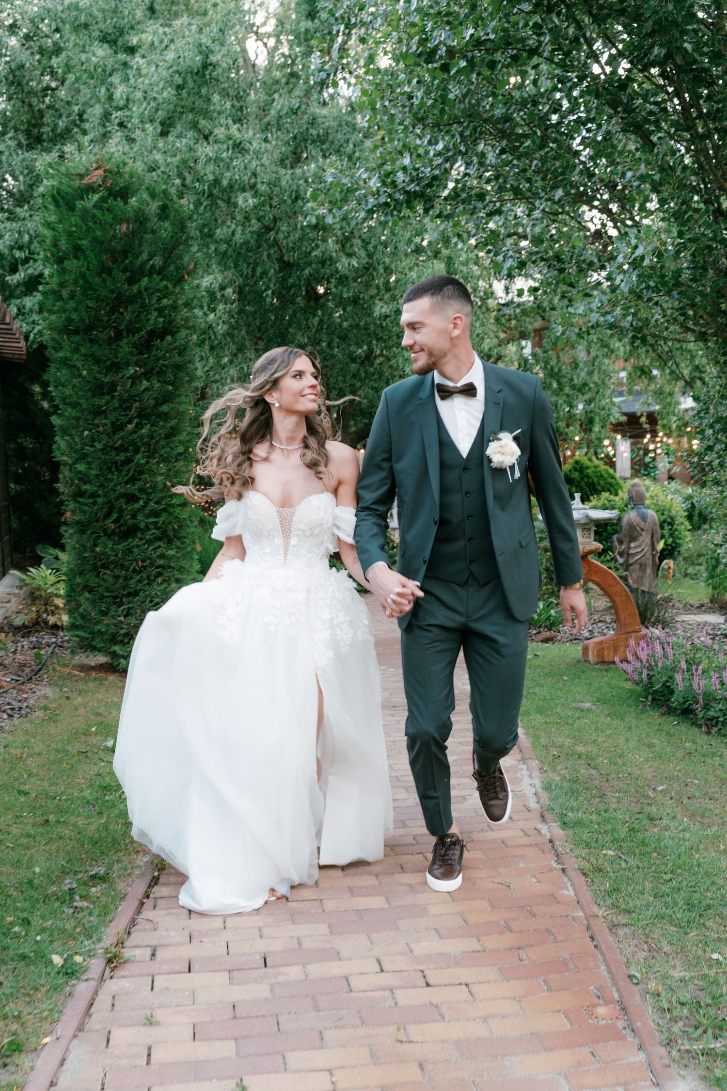 Happy bride and groom holding hands and walking together along brick garden pathway outdoors. Mallorca wedding photographer and filmmaker.