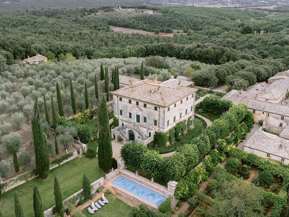 Aerial view of a grand Tuscan villa surrounded by cypress trees, olive groves, and a swimming pool in the Italian countryside.