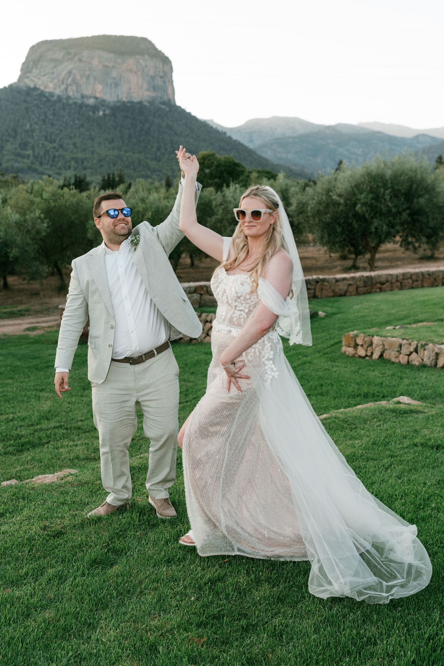 Bride and groom holding hands up in celebration outdoors, with mountain scenery and olive trees behind them.