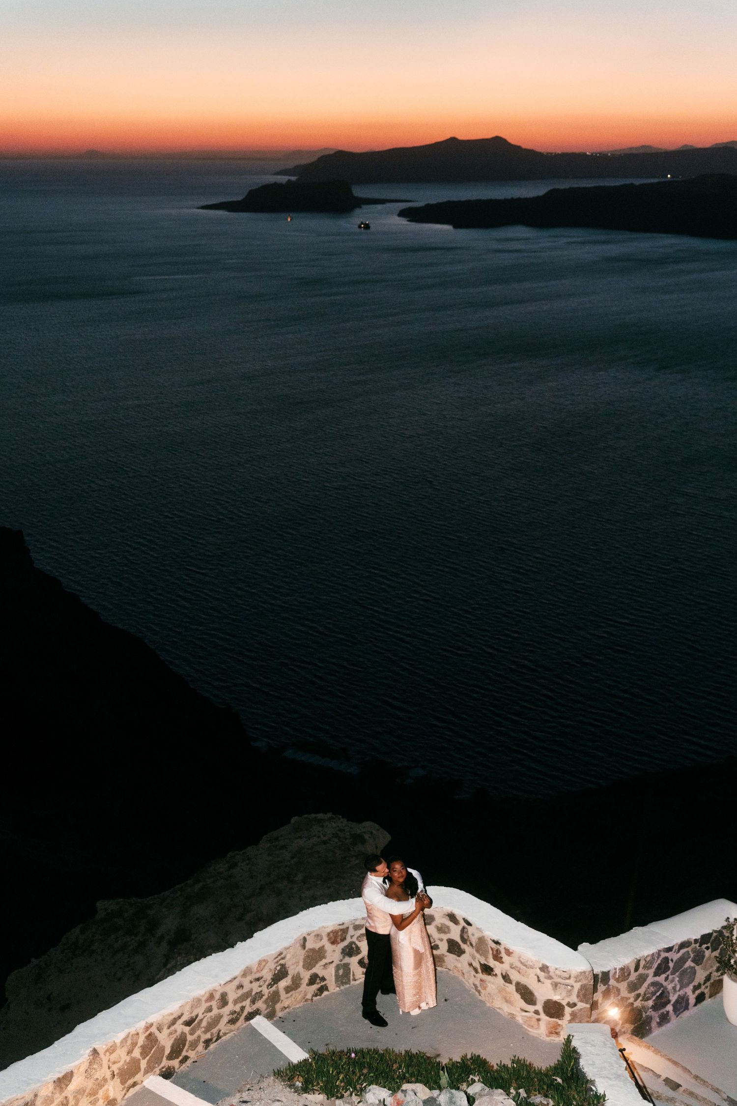 Couple embracing at sunset on a Santorini terrace overlooking the caldera and Aegean Sea at dusk.