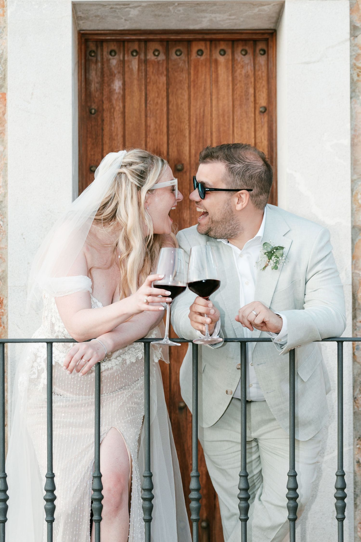 Bride and groom sharing wine on a balcony, smiling at each other in wedding attire on a sunny day in Mallorca.
