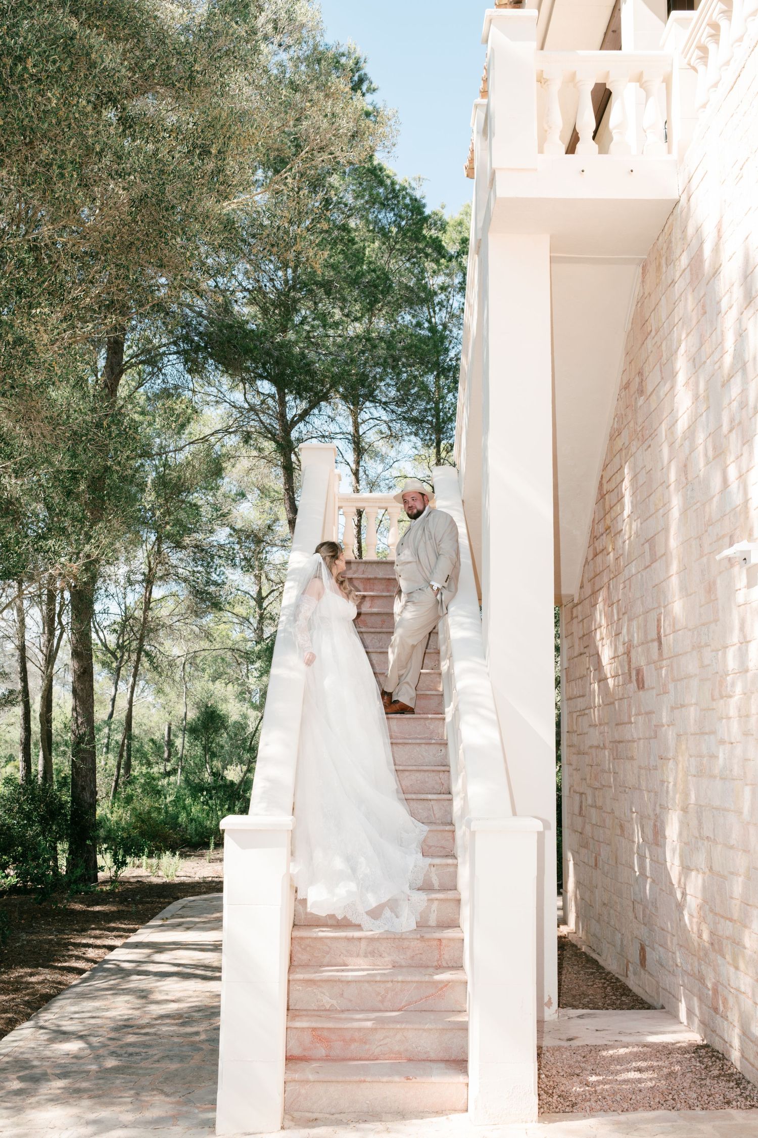 Bride and groom share a romantic moment on outdoor stairs at a elegant wedding venue surrounded by lush trees in Mallorca.