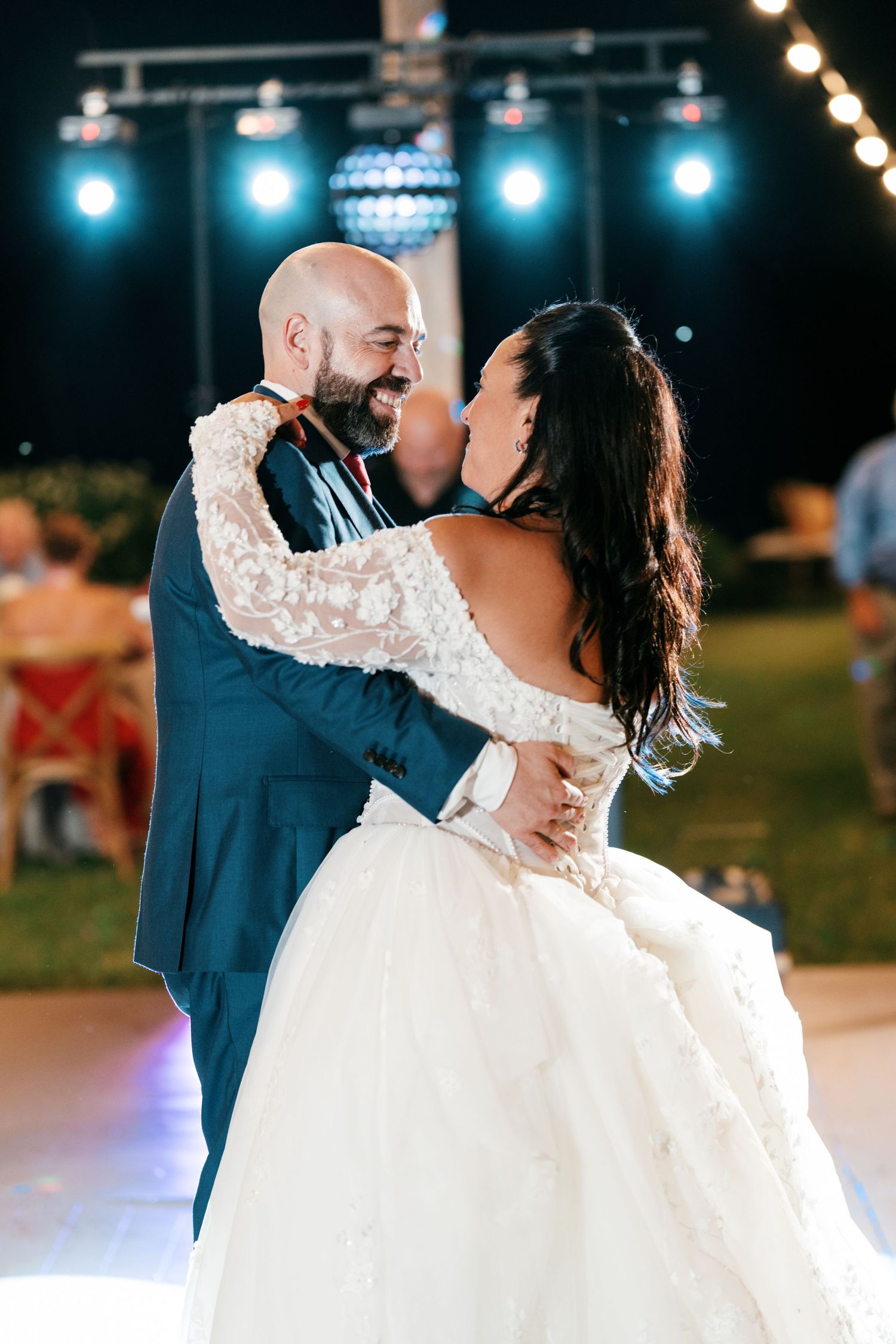 Bearded groom in a blue suit shares a close first dance with his bride in a lace-sleeved gown under blue stage lights.