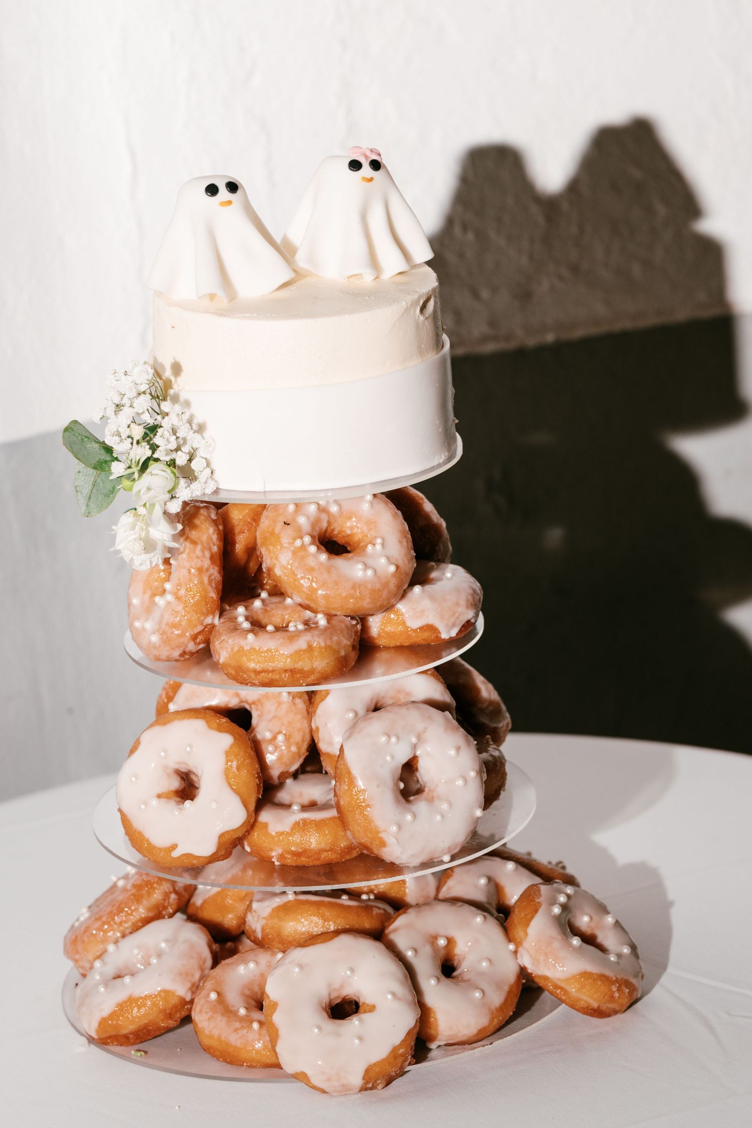A wedding dessert tower of glazed donuts topped with a small white cake and two cute ghost figurines.