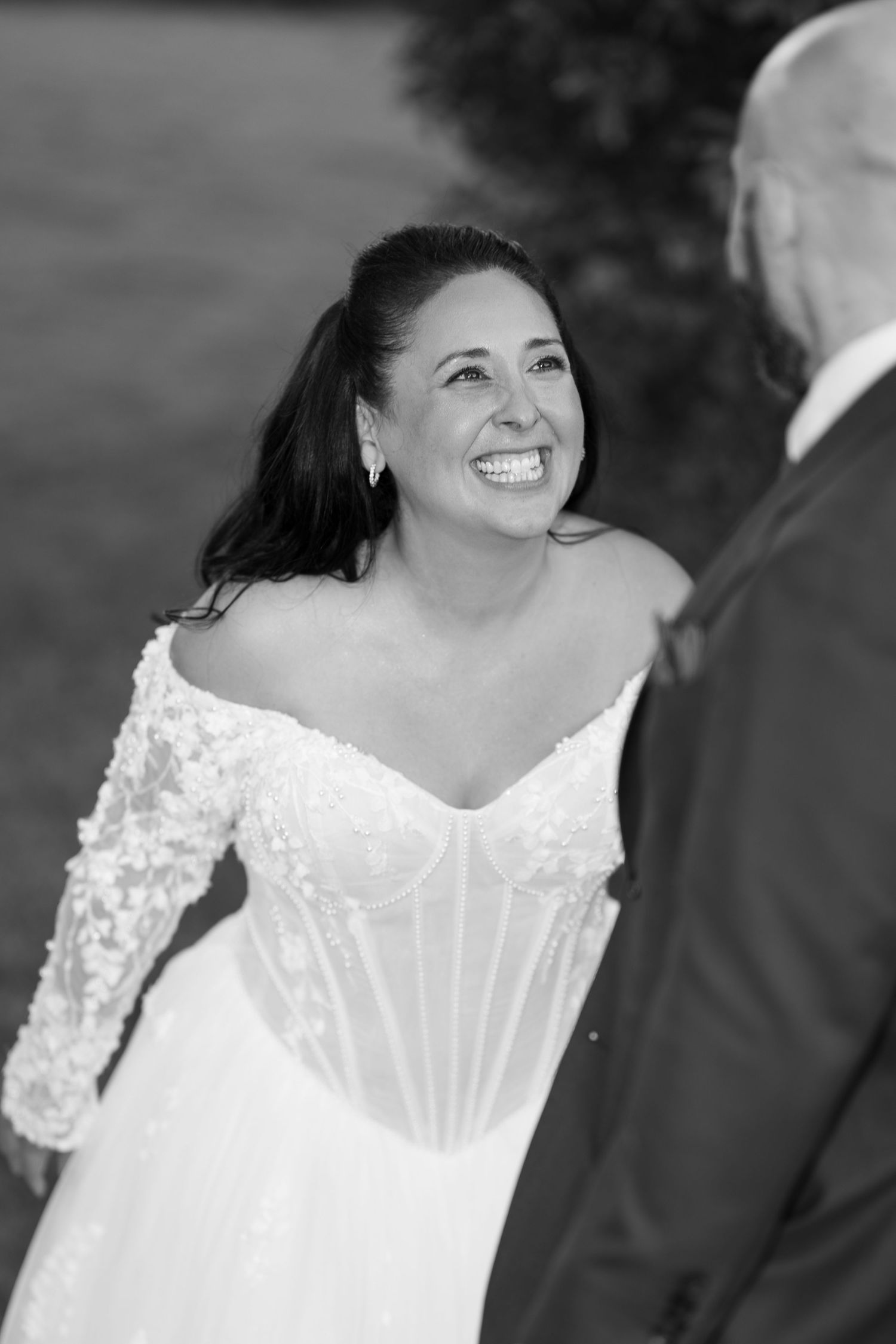 Black and white photo of a smiling bride in an off-shoulder lace wedding gown laughing joyfully while facing her groom outdoors.