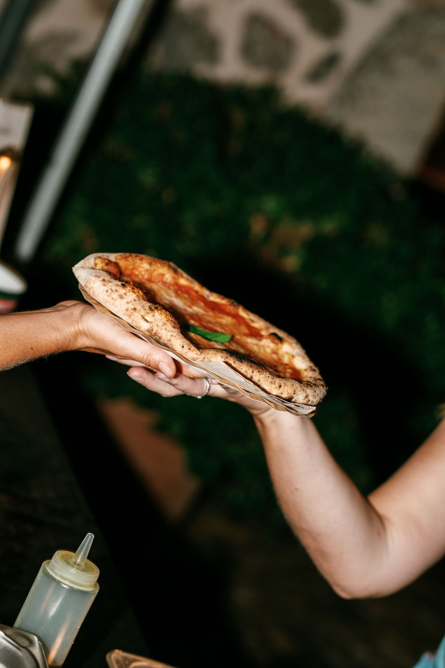 Two hands hold a folded Neapolitan-style pizza with a charred, blistered crust, tomato sauce and fresh basil outdoors.