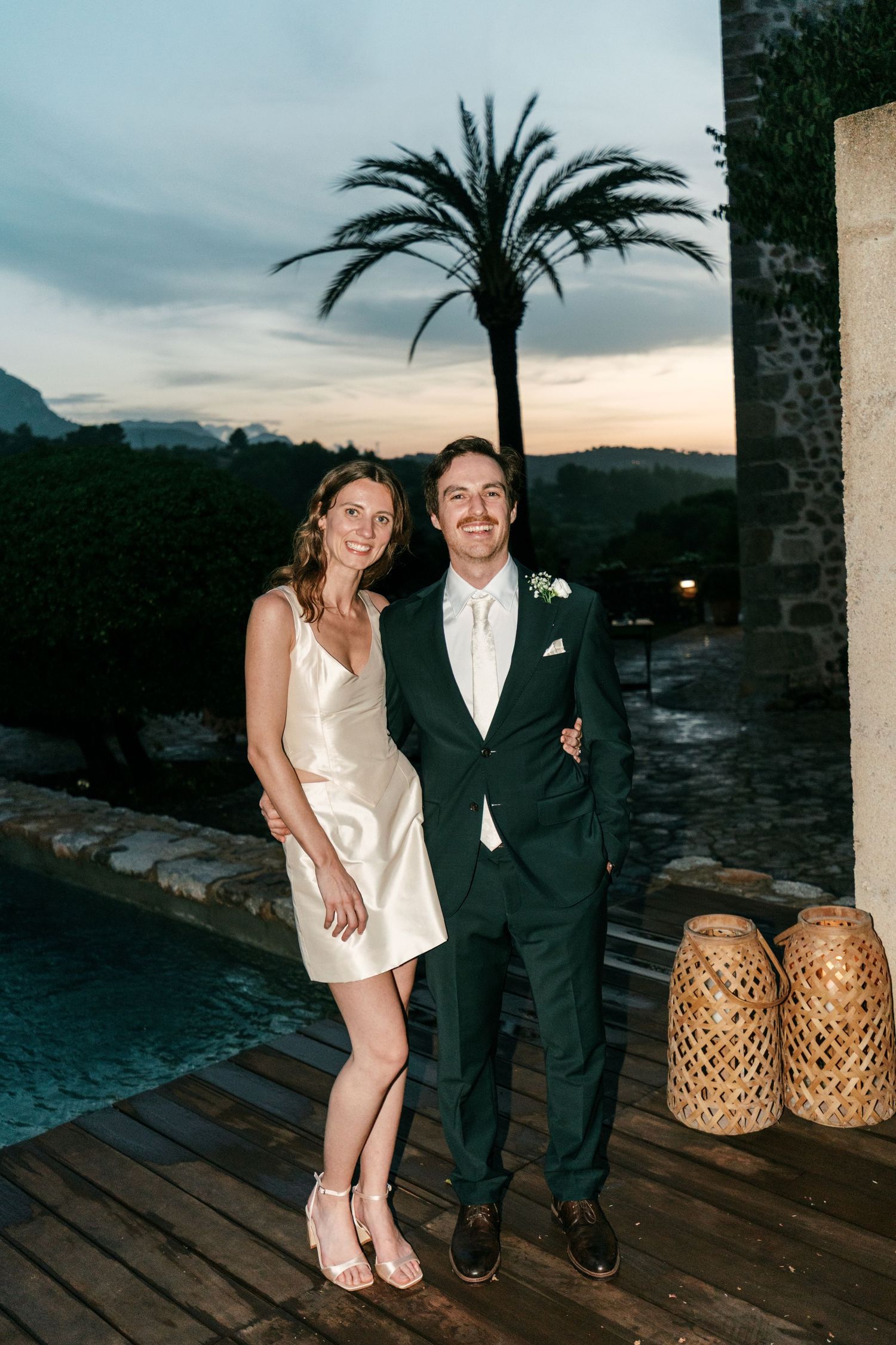 A bride in a satin mini dress and a groom in a green suit pose by a pool with a palm tree at dusk.