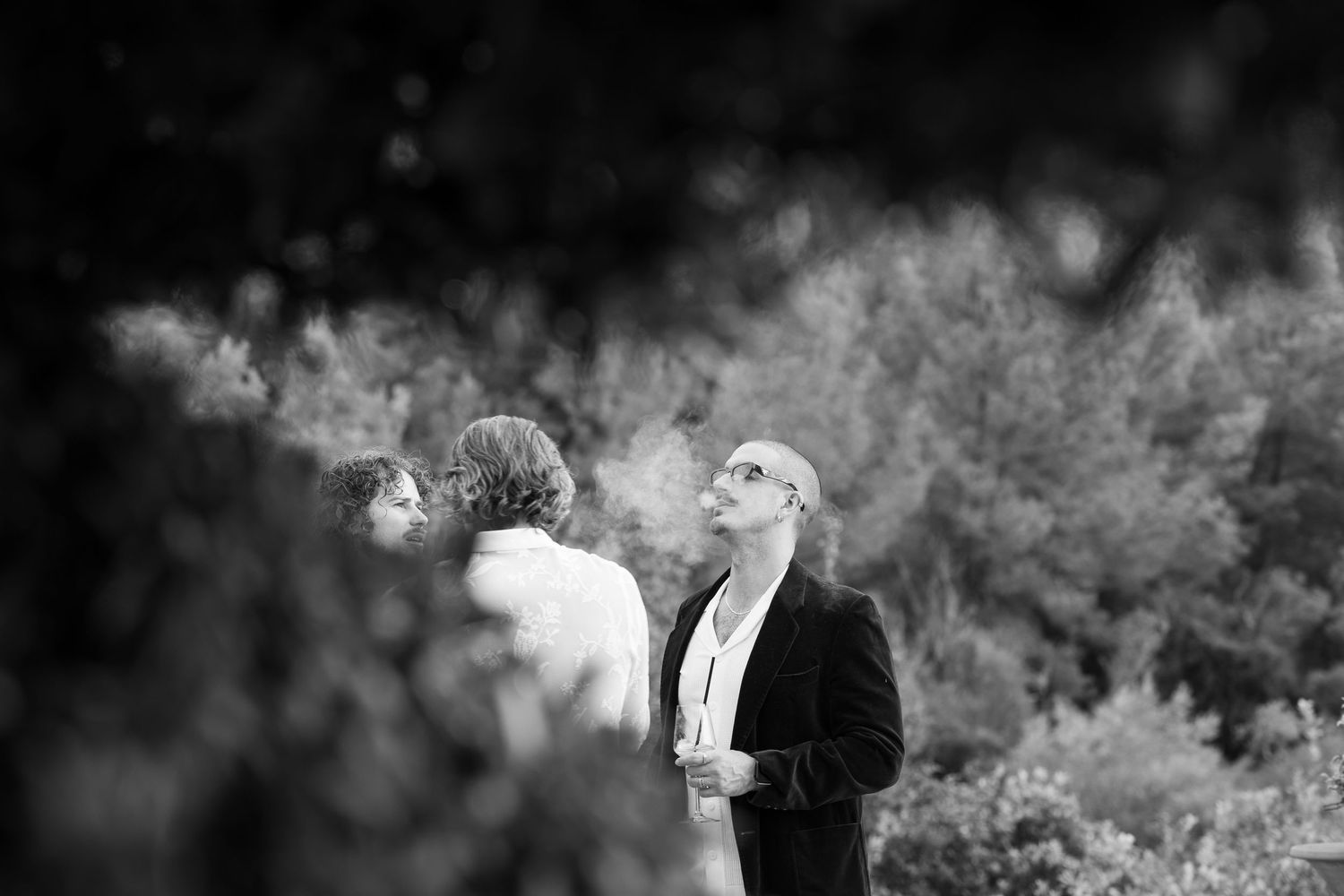 Black and white photo of a bearded man in a dark jacket gazing upward beside two people, framed by blurred leaves and trees.