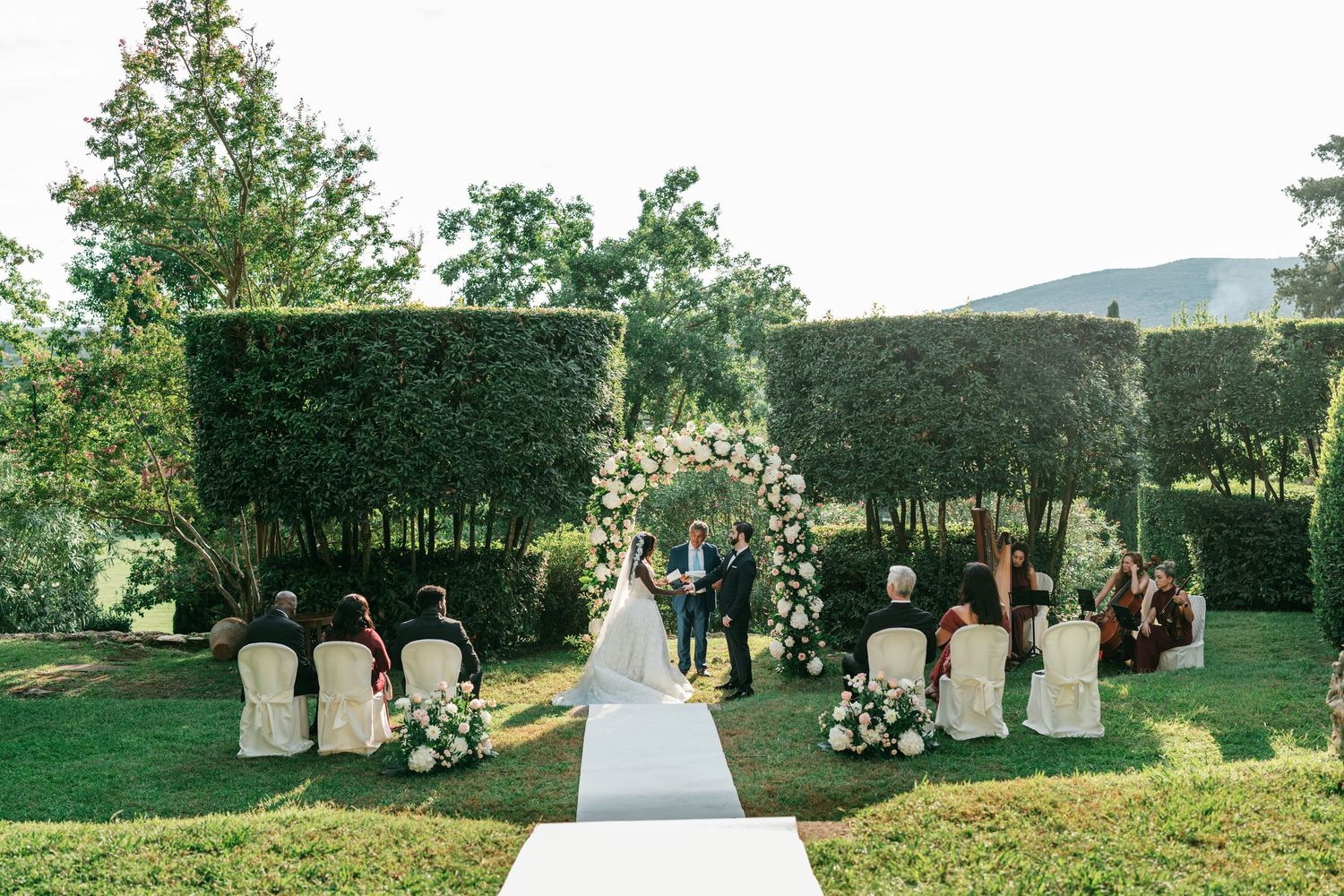Wedding ceremony on a green lawn with guests seated on white chairs as a couple stands beneath a floral arch near tall hedges.
