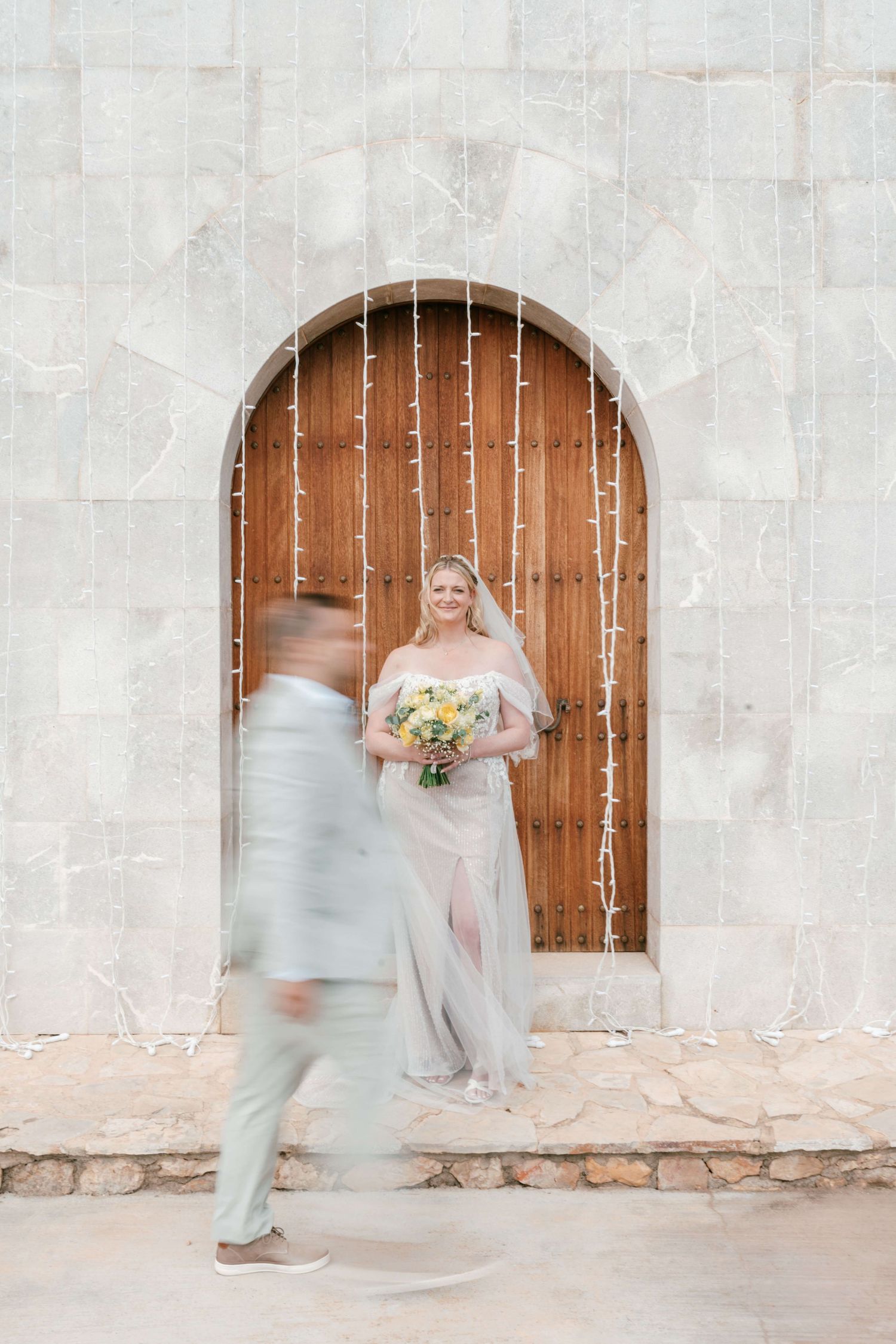 Bride in elegant gown holds bouquet by wooden arched door as groom walks past in light gray suit in Mallorca.