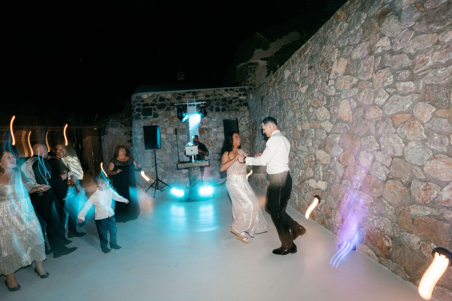 Couple dancing at outdoor wedding reception with guests holding light wands near stone wall at night.