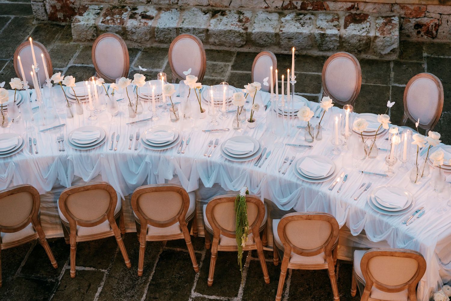 Elegant oval wedding reception table with taper candles, white linens, and rattan chairs on stone floor.
