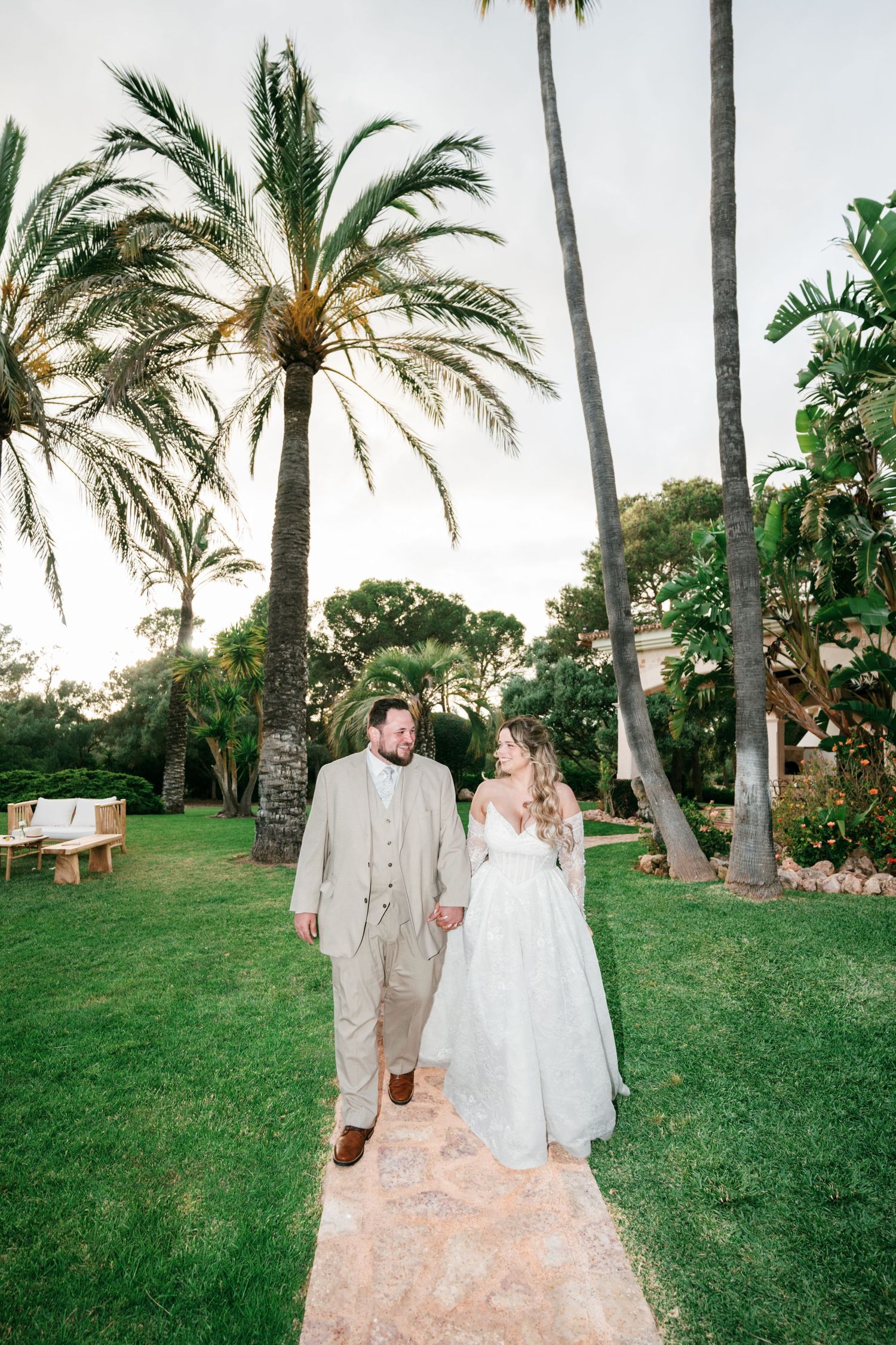 Bride and groom walk hand-in-hand on a garden path surrounded by palm trees at an outdoor wedding venue.
