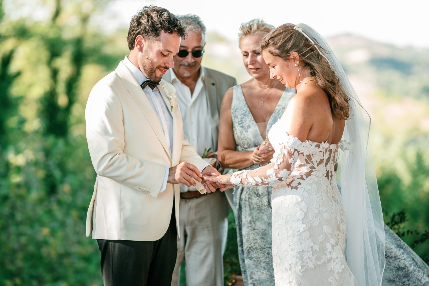 Bride and groom exchanging rings outdoors at a wedding ceremony with guests watching in a lush green setting.