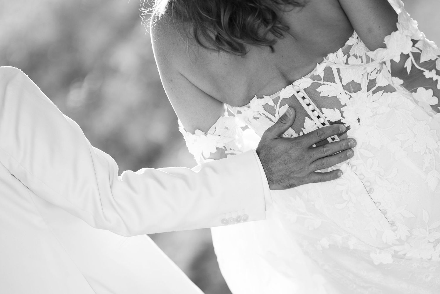 Black and white photo of a groom fastening the back of a bride's lace wedding dress.