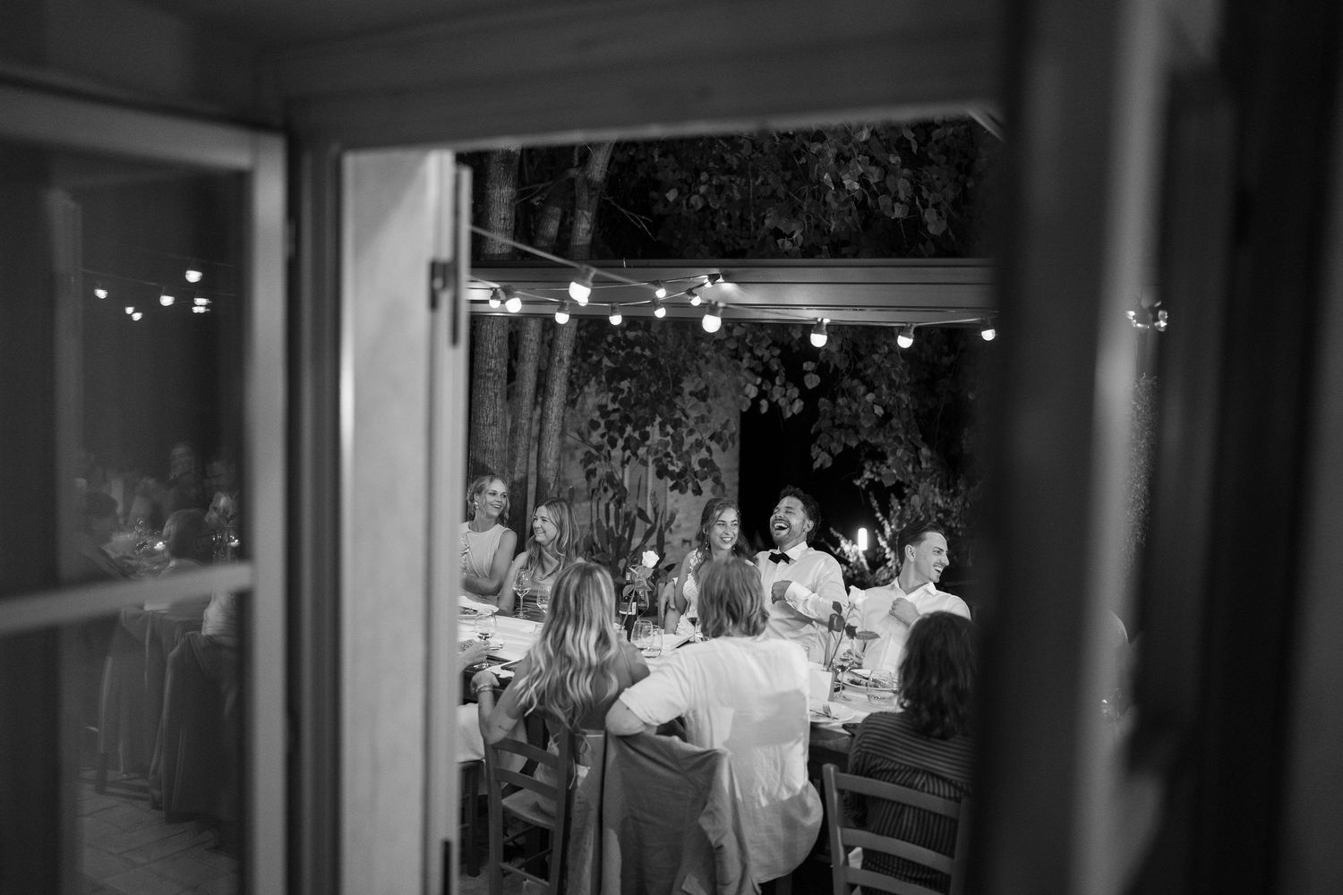 Black and white view of wedding guests dining at outdoor reception through window.