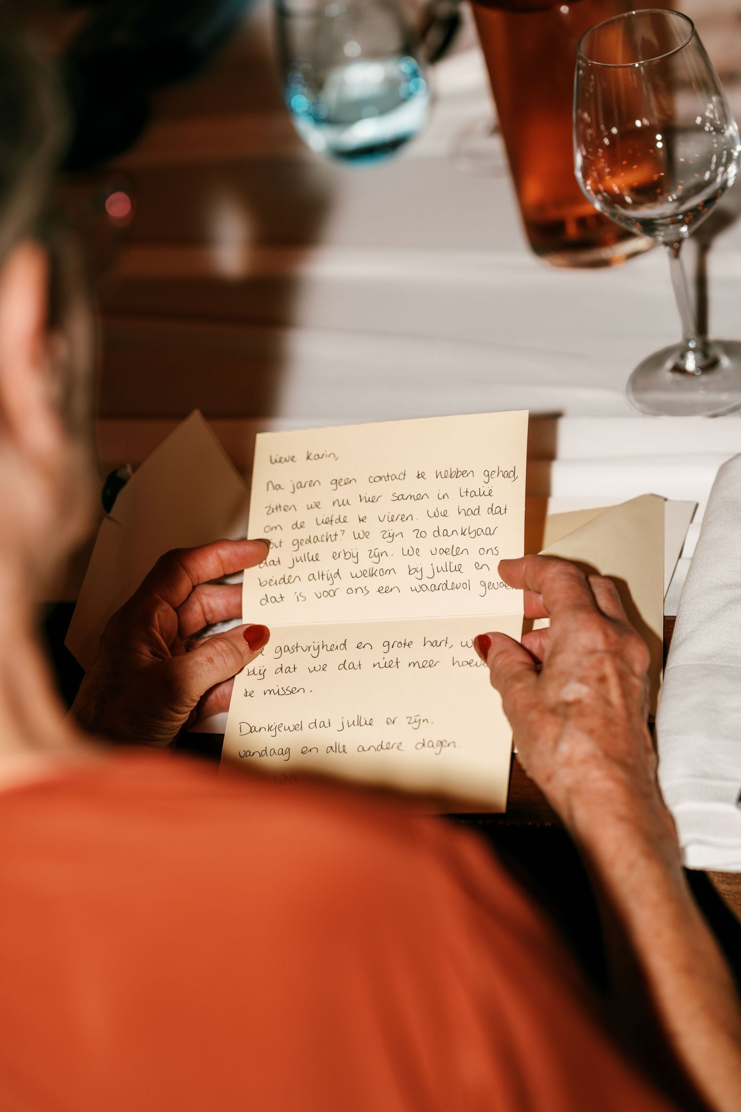 Elderly person reading a handwritten letter at a dining table with wine glasses in the background.