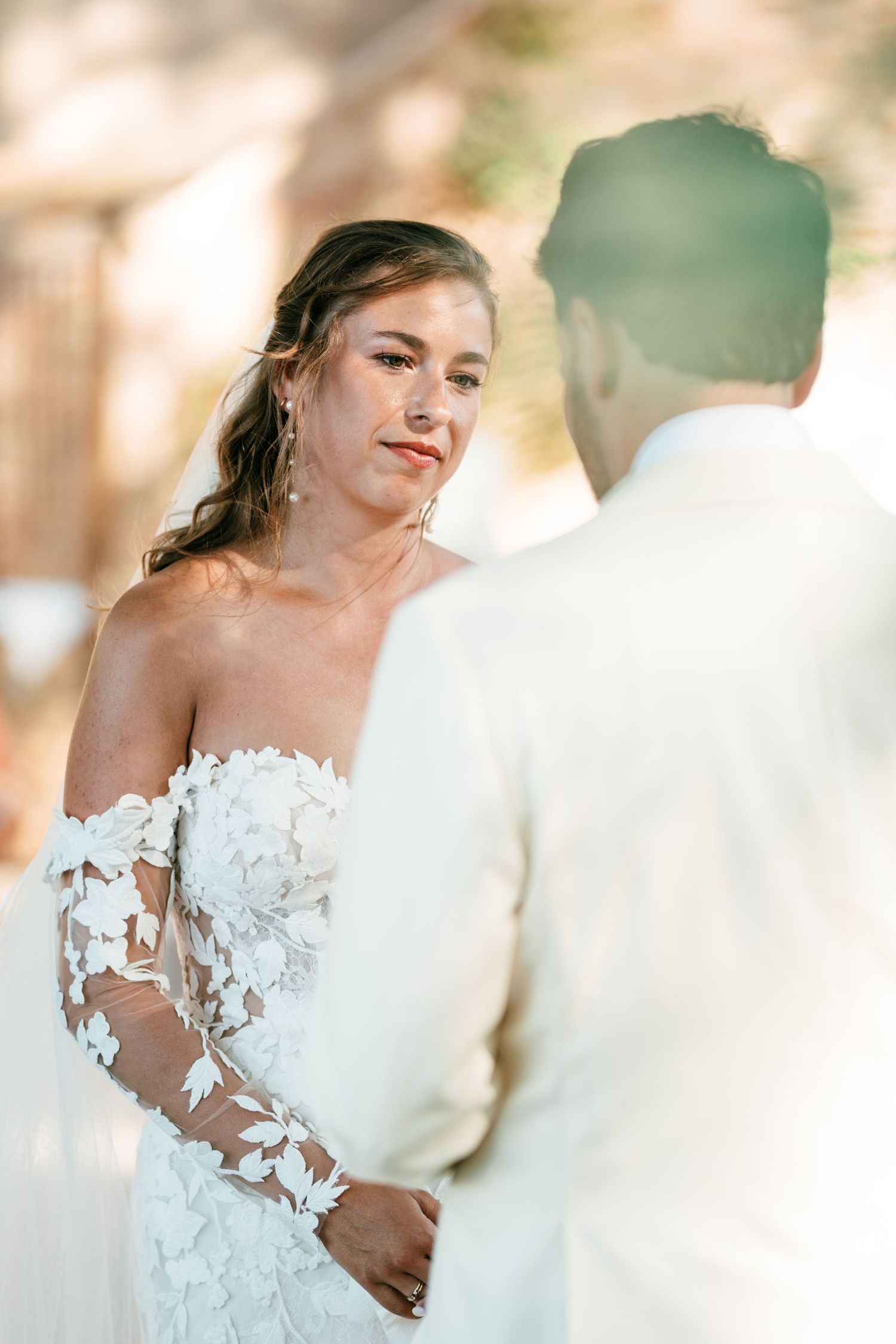 Bride in floral lace off-shoulder gown gazes at groom during outdoor wedding ceremony.