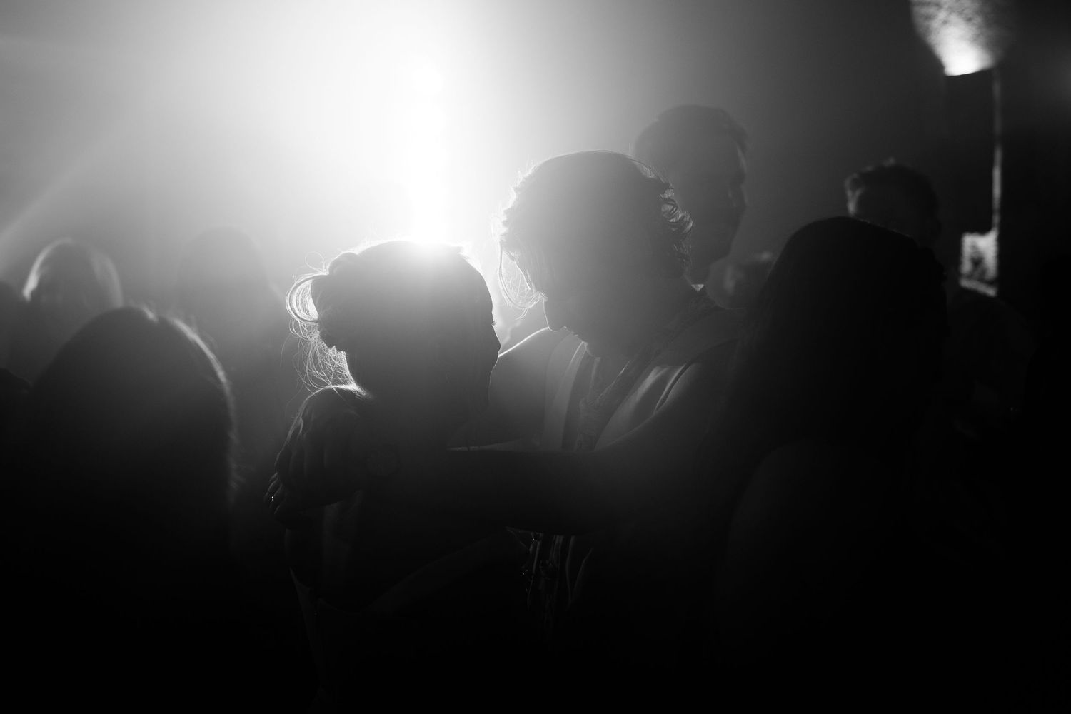 Silhouettes of people dancing in a dimly lit nightclub with bright backlight glowing through haze in a black and white photo.