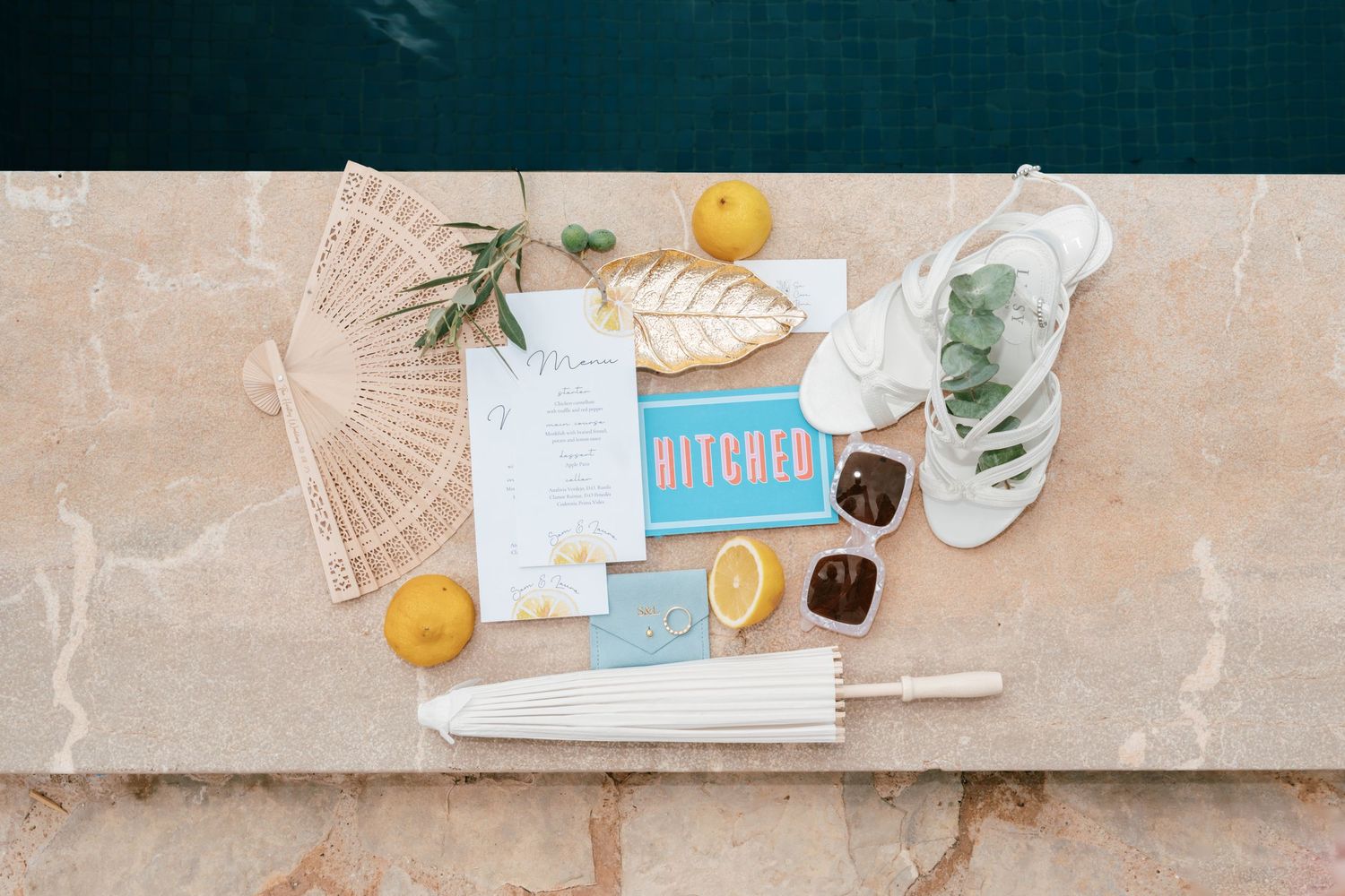 Wedding flat lay with citrus lemons, fan, umbrella, and Hitched stationery by a pool. Mallorca wedding photographer, filmmaker