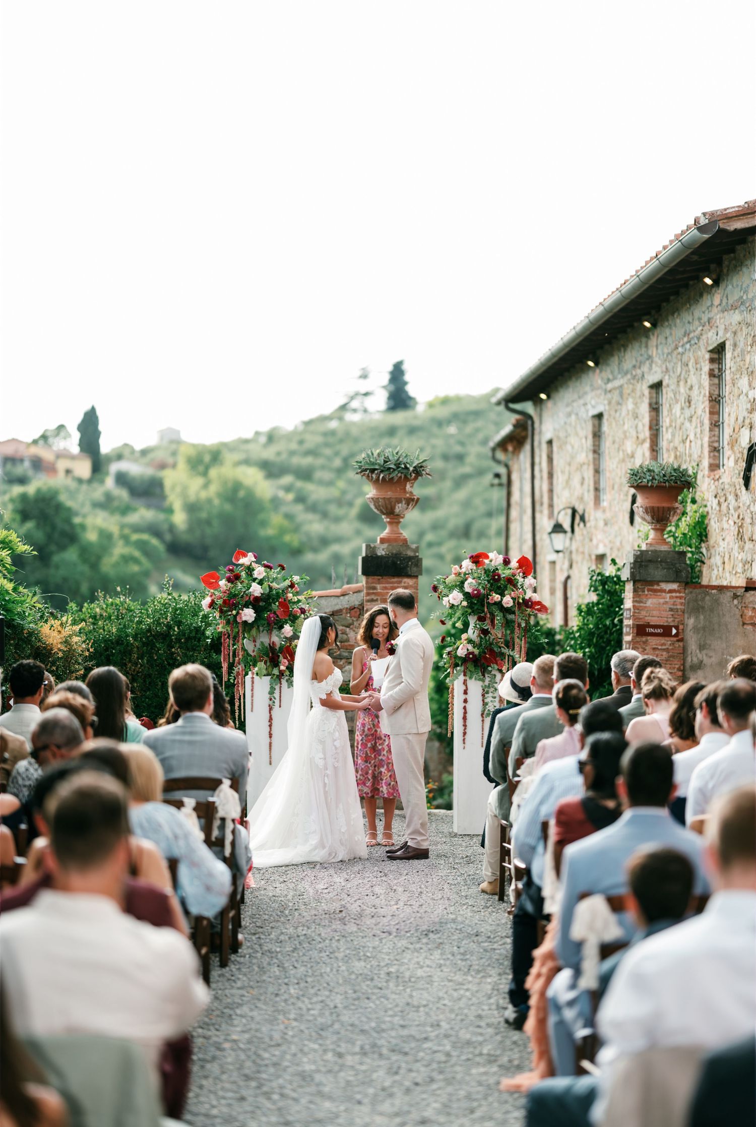 Bride and groom exchange vows at an outdoor Tuscan wedding ceremony as guests watch beside red floral arrangements and stone urns.